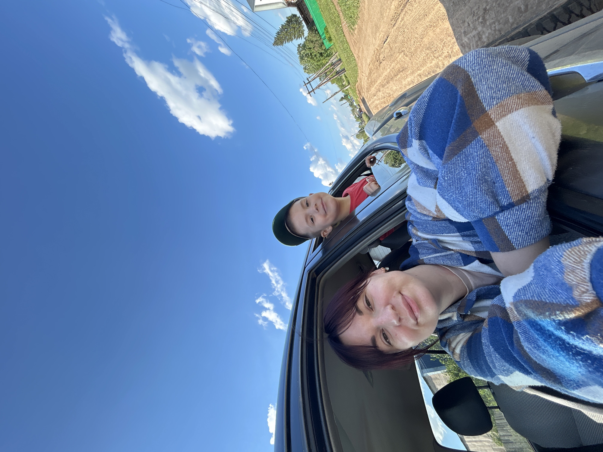 In a selfie taken from inside a dark-colored car, a young woman with dark hair and a plaid shirt smiles at the camera. She is leaning on the open car door, with her arm visible. Behind her and to her right, a young boy wearing a red shirt and a green cap is looking out of the car window and also smiling. The car is on a dirt road, likely in a rural area. The background shows a bright blue sky with scattered white clouds, green trees, a wooden fence, and some utility poles. The scene appears to be set in the town of Foky, Russia, given the context of the image. The overall impression is one of a casual outing or road trip on a sunny day.