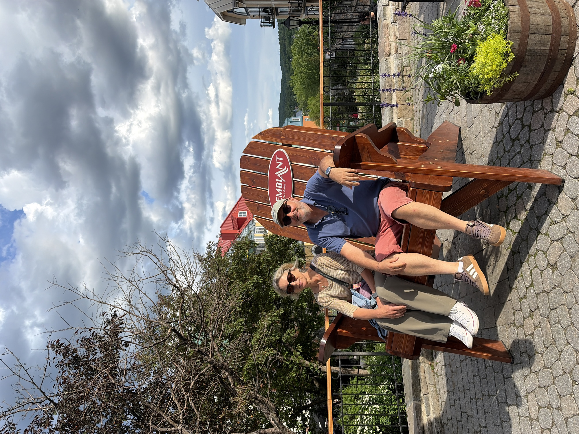A middle-aged couple, a woman with blonde hair and sunglasses, and a man with a cap and sunglasses, are seated on a giant wooden Adirondack chair. The woman is wearing a beige knitted sweater and olive-colored pants, while the man is in a blue polo shirt and salmon-colored shorts. They are positioned on a cobblestone patio with a backdrop of trees, buildings with red roofs, and a cloudy sky. A barrel planter filled with various flowers and greenery is located to the right of the chair. The chair has a red oval logo with white text that reads "TREMBLANT" and "Mont-Tremblant, Quebec". This scene is in the pedestrian village of Mont-Tremblant, Quebec, Canada.