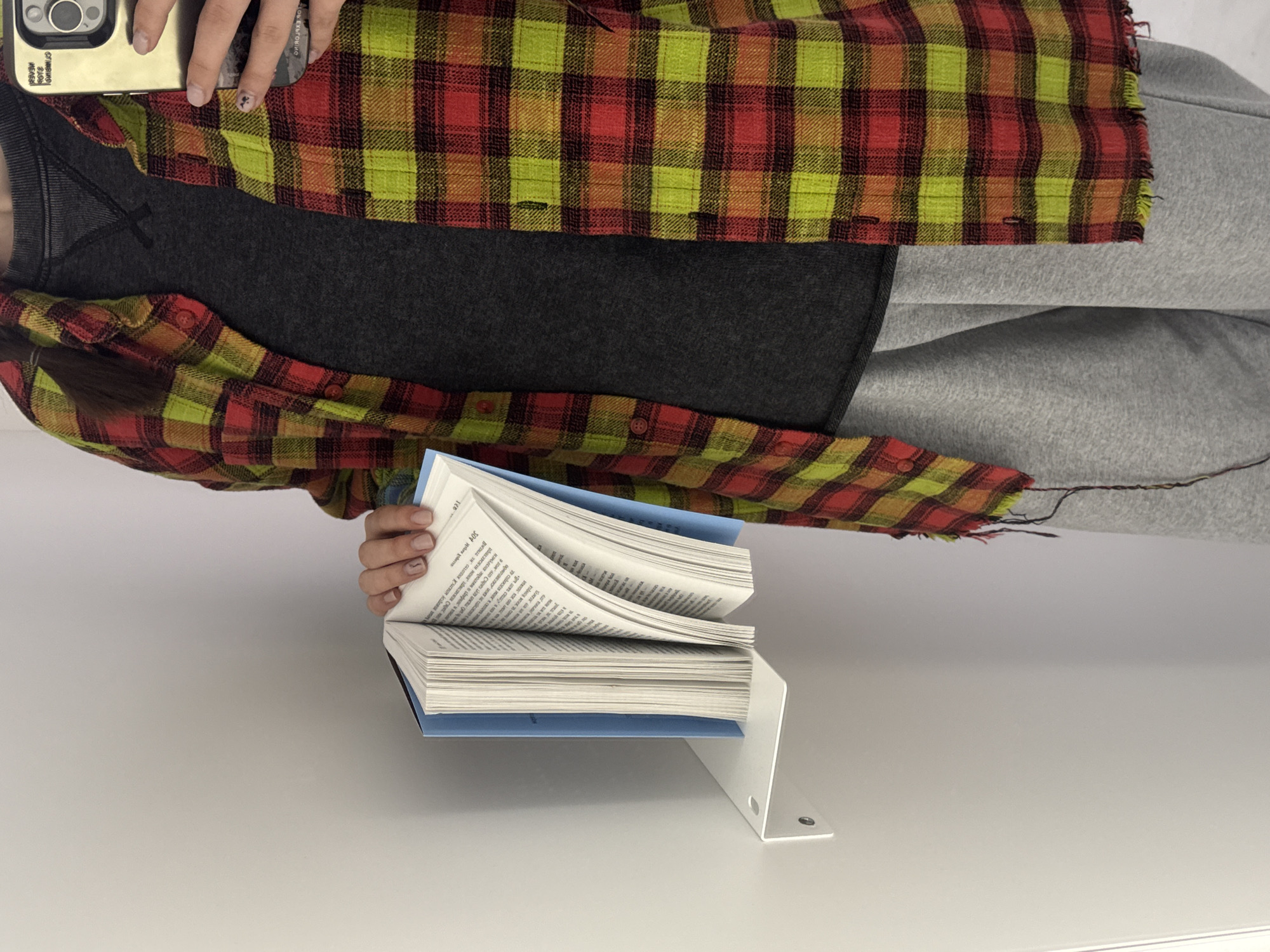 The image depicts a person holding an open book on a white metal stand against a white background. The person is wearing a gray top and gray sweatpants, with a red and yellow plaid shirt over the top. Their left hand is visible holding the book open, and their right hand is holding a mobile phone near their head. The book has blue cover boards and the pages are filled with text. The white metal book stand is minimalist in design with a right angle. The image is brightly lit and appears to be taken in an indoor setting.