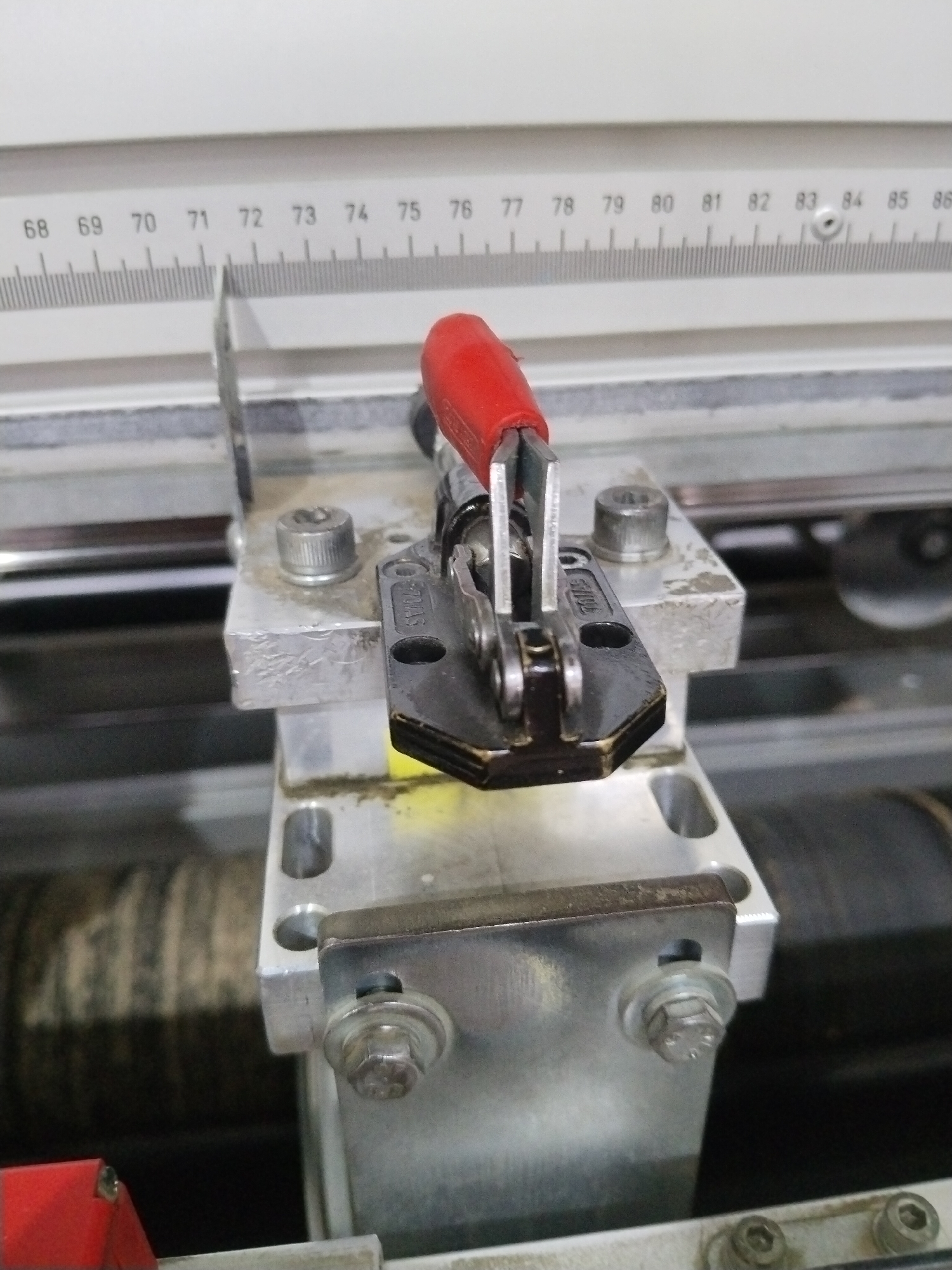 The image shows a close-up, eye-level view of a clamp mechanism attached to a piece of industrial equipment. The clamp, which has a red handle, is made of metal and is secured to a silver-colored bracket by bolts. A ruler with markings from 68 to 86 is visible at the top of the image, providing a scale for the equipment. The background is out of focus, showing parts of the machinery and possibly a textured surface. The overall impression is one of a functional industrial setting. The image does not contain information that would allow for localization to the city of Гожскі сельскі Савет, Belarus, or any other specific geographic location.