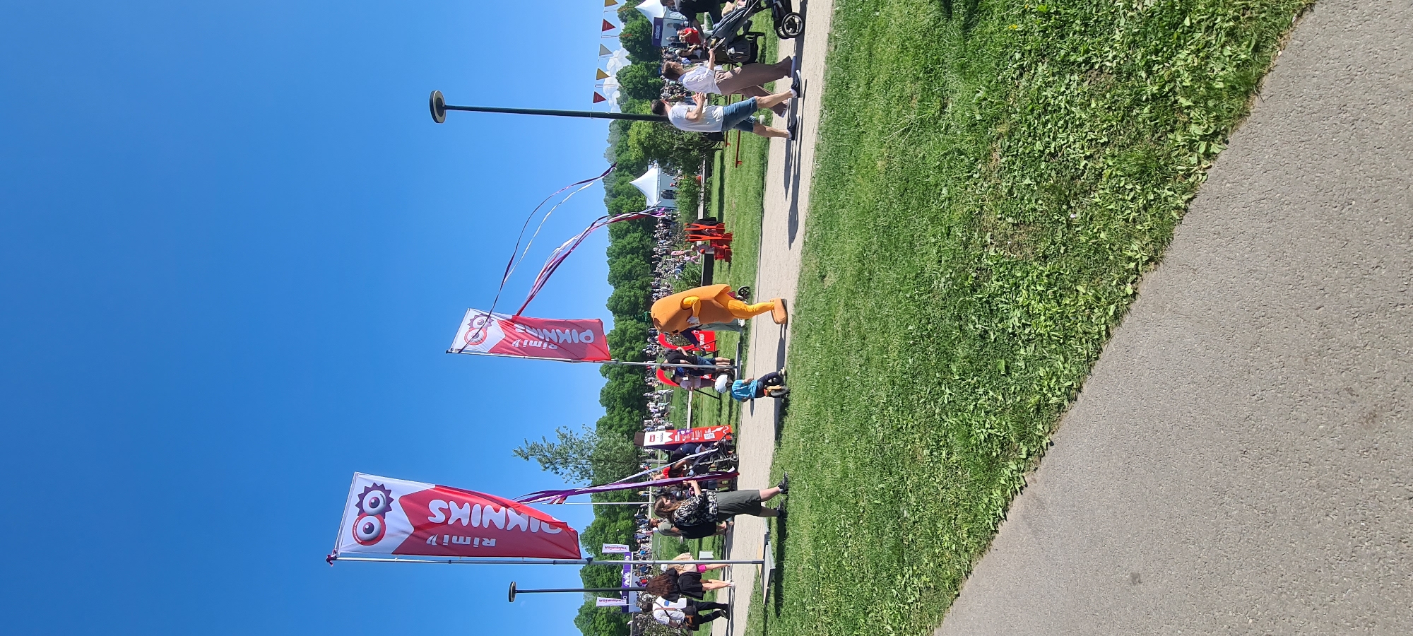 The image depicts an outdoor scene in Riga, Latvia, showcasing a Rimī Piknik festival. A wide crowd of people gathers in a grassy area under a clear blue sky. The foreground is dominated by a paved pathway, adjacent to the grass. Several vertical banners displaying the "Rimī Piknik" logo are aligned along the pathway. Some of the banners have decorative streamers. A person in a large orange mascot costume stands near one of the banners. Pedestrians are strolling along the pathway. In the background, there's a cluster of white tents and a number of colorful triangular pennants.