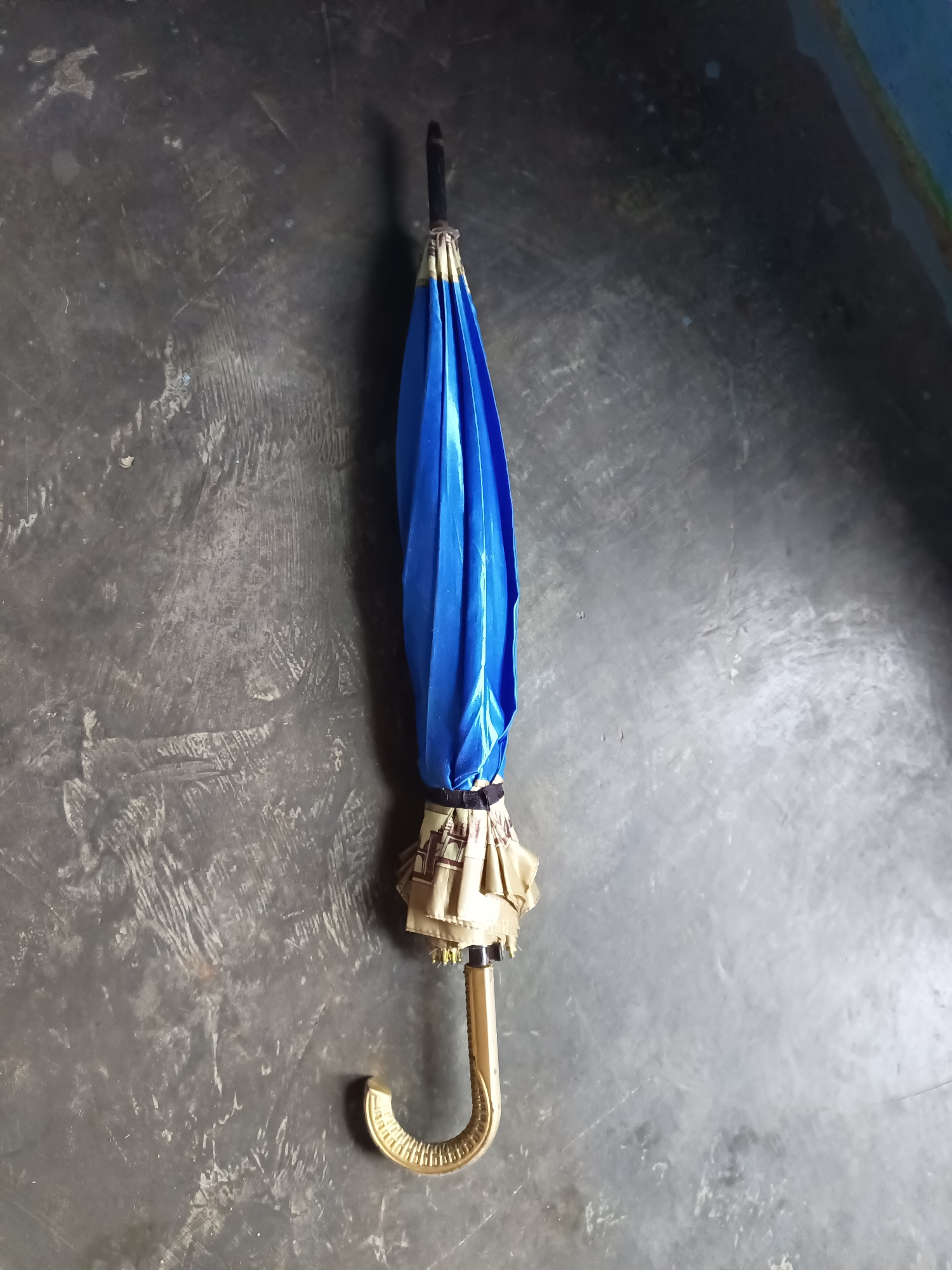 A closed blue umbrella with a golden hook handle rests on a textured gray surface. The umbrella is positioned vertically in the center of the frame, with the handle at the bottom and the folded fabric at the top. The surface it lies on appears to be a floor or ground with visible scuffs and variations in texture and color. The lighting suggests an indoor setting or an overcast day. This image was taken in Terbanggi Marga, Indonesia.