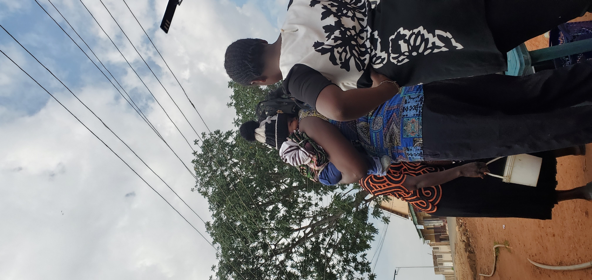 In Gura Top, Nigeria, three individuals are visible in an outdoor setting under a cloudy sky with power lines overhead. A woman on the left, wearing a black skirt and a patterned orange and black top, holds a white bucket in her left hand. In the center, a woman is holding a baby wrapped in a striped cloth and wearing a black, white, and grey beanie. The baby appears to be asleep. The woman holding the baby is wearing a blue, black, and patterned garment. To the right, another woman, seen from the back, wears a black and white patterned top. Behind the people, a large tree with green leaves is prominent. In the background, there are buildings and a road, with a light pole visible on the right side of the frame. The ground is composed of reddish-brown dirt.