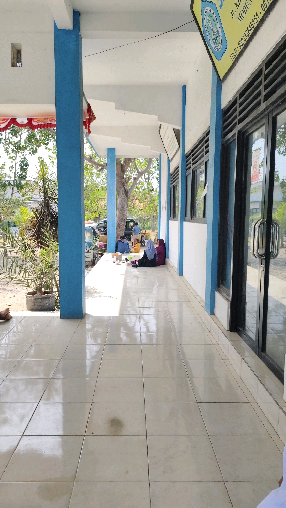 This image captures a covered walkway in front of a building with blue and white trim. The walkway is paved with cream-colored tiles that reflect the sunlight, creating a bright and slightly glossy appearance. To the left, a large blue pillar stands prominently. Beyond the pillar, there are trees and some parked vehicles. A few individuals are seated on the walkway. Two women wearing hijabs are sitting on the ground, facing away from the camera, with one appearing to be looking at something on the ground. Another person is seated to their left, and a fourth person is standing in the distance near the parked vehicles. There are also some small items scattered on the ground near the seated individuals, possibly food containers or personal belongings. A sign with Indonesian text and a logo is visible on the upper right side of the building. The overall scene suggests a relaxed atmosphere, possibly a break or informal gathering area outside a school or community building in Pangpajung, Indonesia.