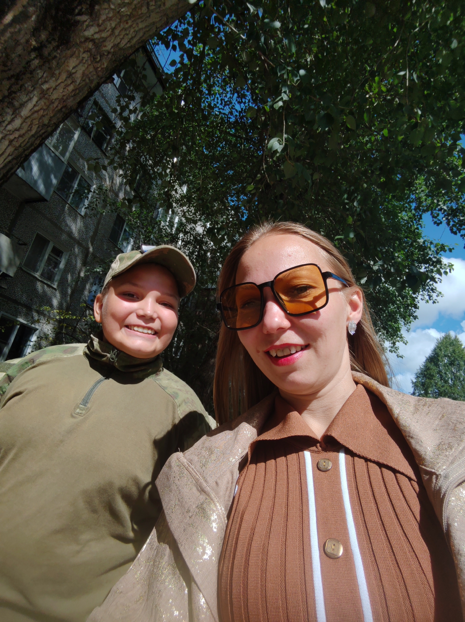 Two individuals are captured in a close-up, low-angle selfie outdoors. The person on the left is wearing a camouflage baseball cap and a green zippered shirt with a high collar. They are smiling directly at the camera. The person on the right, also smiling, wears large, orange-tinted square sunglasses, a brown collared shirt with white vertical stripes and gold buttons, and a shimmering gold jacket. They also have a prominent silver earring. Behind them, a residential building with multiple windows is visible, partially obscured by the dark bark of a tree trunk on the left and lush green foliage. The sky above is bright blue with scattered white clouds. The scene suggests an outdoor setting in a city environment. Given the style of the building and the overall atmosphere, this location could be in Syktyvkar, Russia.