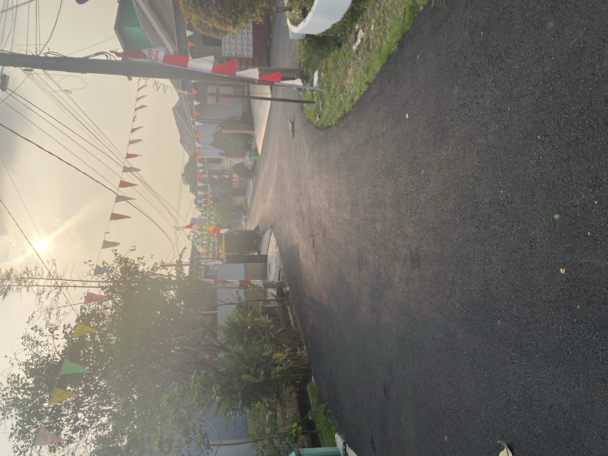 The image depicts a street scene in Depok, Indonesia, characterized by the festive decorations of triangular flags strung across the road and the presence of Indonesian flags. The flags, red and white, are a prominent visual element, with some affixed to poles and others strung along wires. The road is paved and curves to the right. On either side of the street, residential houses with tiled roofs are visible. Green foliage and trees line the sides of the road, and some grass is present. The sky is bright with the sun, suggesting it is daytime, possibly late afternoon given the warm, hazy light and the long shadows. There are no people or specific actions clearly visible in the image, creating a static depiction of the environment.