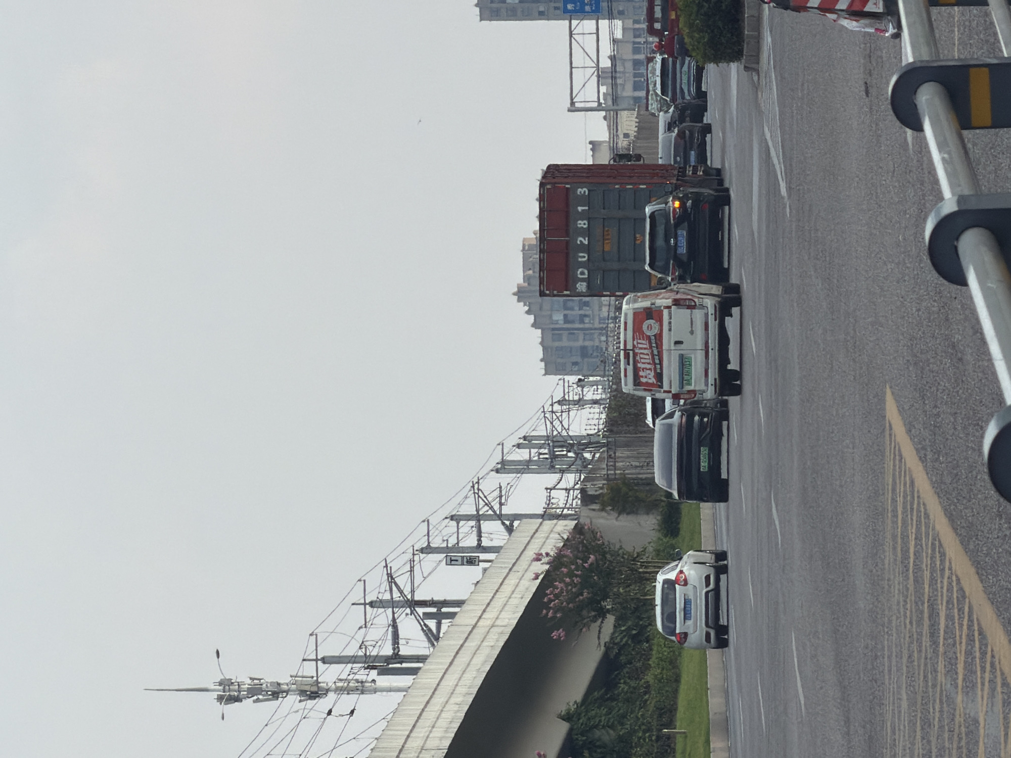 The image depicts a street scene in the city of Jiangjin, China, with a focus on a traffic jam. Multiple vehicles, including cars, a white van with red lettering, and a large red truck with a grey container, are stopped on a multi-lane asphalt road. The van has a license plate reading "A AH7037." A black SUV behind the van has a license plate that appears to read "LU CQ678." The truck's container has "DU 2813" visible. The road is marked with yellow and white lines, and some traffic cones are visible in the foreground. Elevated railway tracks with power lines stretch across the background, supported by numerous poles. A concrete overpass runs along the left side of the image, with green foliage and a flowering shrub growing beneath it. Buildings, some appearing to be residential or office blocks, are visible in the distance. The sky is overcast and pale.