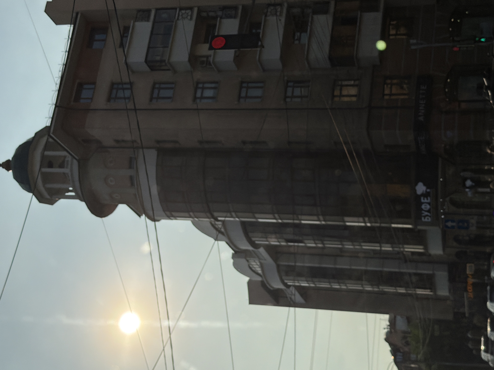 The image is a street-level view of a multi-story building in Krasnoyarsk, Russia, captured during daylight. The building features a rounded section with a dome-like structure on top and a glass-paneled facade on the lower levels. Overhead power lines crisscross the sky. A traffic light with a red signal is visible on the right side of the building. Signs on the building are partially obscured, but "БУФЕ" (Buffer) and "ANNETTE" are discernible. The sun is bright and partially obscured by atmospheric haze or lens flare, casting a strong glare on the left side of the image. The lower portion of the image shows street-level activity with what appear to be vehicles and pedestrians, though these are not clearly detailed.