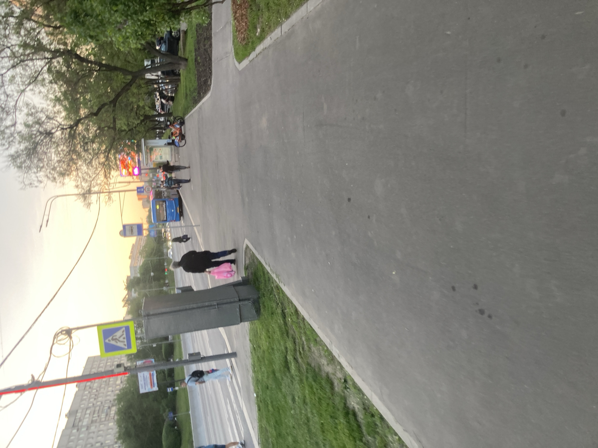 The image depicts a street scene in Moscow, Russia, viewed from a slightly elevated perspective. A pedestrian walkway and a road intersect at a crossroads. A person carrying a pink bag is crossing the road. Other pedestrians can be seen in the distance. A bus and several cars are on the road. There are trees and buildings visible, suggesting an urban environment. A pedestrian crossing sign is visible. The sky suggests it is either sunrise or sunset.