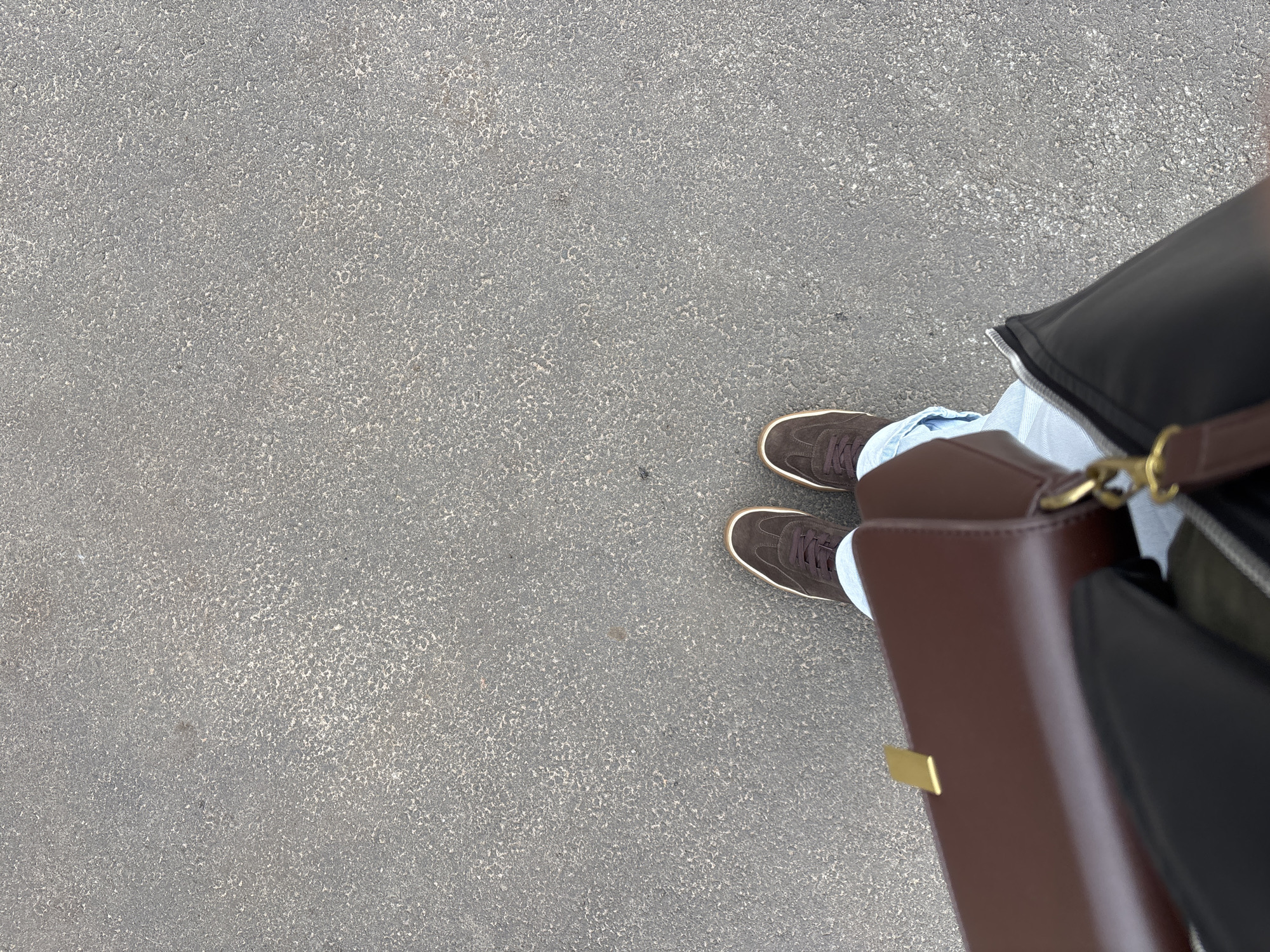 This is a top-down, medium shot of a person's legs and feet standing on a textured gray asphalt surface. The person is wearing dark brown suede athletic shoes with white trim and light blue jeans. A dark leather jacket is visible on the upper right side of the frame, partially obscuring the person's legs. To the right, a structured brown leather handbag with gold hardware is held, appearing to be carried by the person. The scene is likely outdoors, as suggested by the asphalt ground and the casual attire. The lighting appears to be natural, suggesting daytime, but the specific time of day and weather conditions are not clearly discernible. There is no visible text in the image. The location context is Заволжье, Russia.