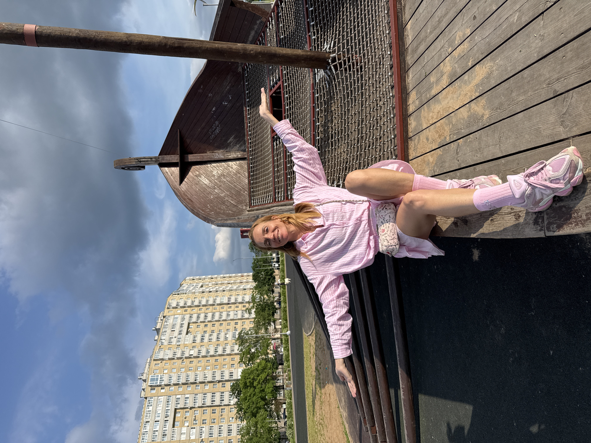 A Caucasian woman with blonde hair is sitting on a wooden surface in front of a large, wooden, ship-like structure. She is wearing a pink striped button-up shirt, a pink skirt, pink socks, and pink sneakers. She has a small, patterned purse slung across her body. Her arms are outstretched, and she is smiling at the camera. The ship structure appears to be a playground feature, with a netting material and wooden supports visible. In the background, a tall, yellow apartment building and green trees are visible, along with a cloudy blue sky. The ground in front of the wooden surface is dark and appears to be asphalt. This scene is located in Kirishi, Russia, as evidenced by the distinctive architecture of the apartment building.