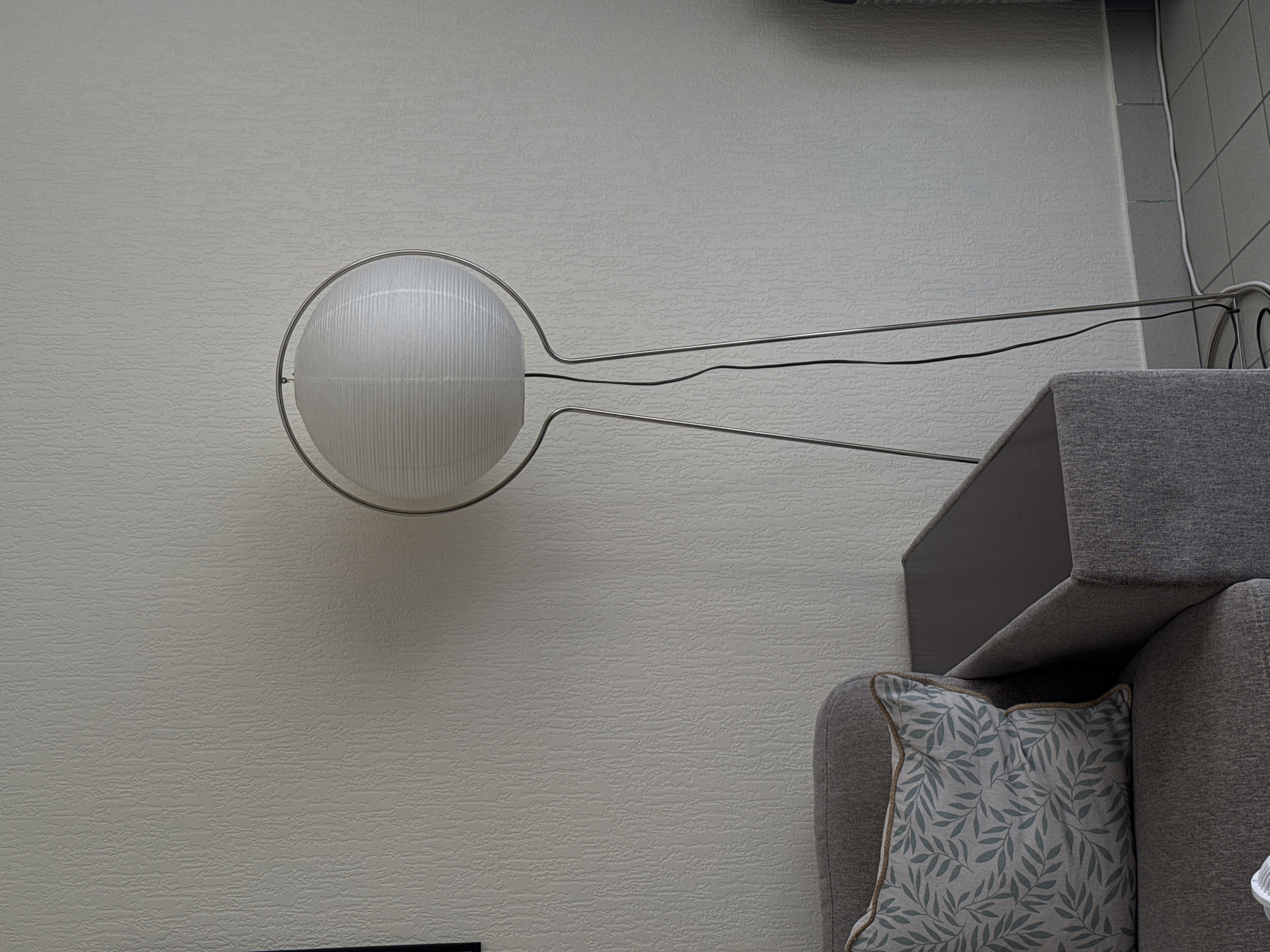 The image depicts an interior scene in a residential setting, possibly a living room, in Vienne, France. On the left side of the frame, a portion of a gray sofa is visible with a patterned throw pillow featuring green leaves. To the right of the sofa, a modern floor lamp with a white, spherical shade and a silver metal frame stands tall. The lamp's base is near the floor, and a black cord extends from it. The background consists of a textured, light-colored wall. The floor appears to be tiled in a gray, grid pattern. The scene is lit, likely by ambient light or the floor lamp itself.