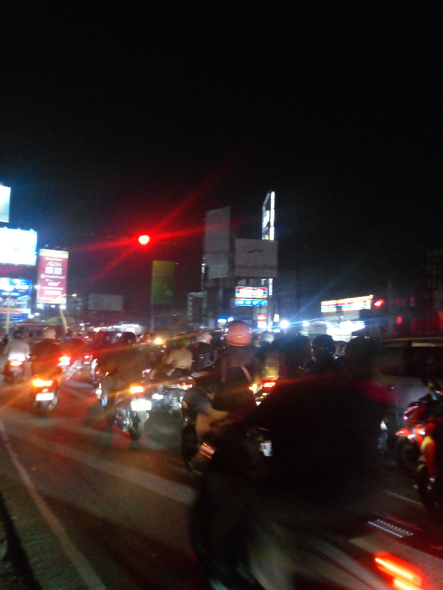 The image depicts a traffic scene at night in the city of Bitung Jaya, Indonesia. A traffic light, illuminated in red, is visible in the upper center of the frame, suggesting a stopped or slowing traffic flow. Numerous motorcycles are present, with their headlights and taillights creating streaks of light due to the long exposure. Some riders are wearing helmets. Billboards and signs, some with illuminated text, are scattered throughout the background, indicating an urban commercial area. The overall scene is dark, with artificial lights from vehicles and buildings dominating the illumination. The motion blur suggests the photograph was taken from a moving vehicle or during a moment of significant movement.