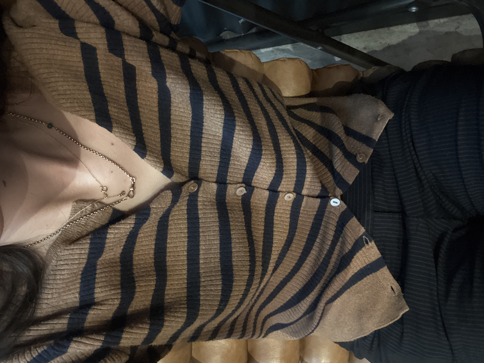 The image presents a close-up, angled view of a person wearing a brown and black striped cardigan-style sweater with buttons partially fastened. A gold necklace is visible against the skin. The person is sitting on a tan cushioned surface. Beside the person, there is a black fabric-like item. The setting is not explicitly revealed in the image, making it impossible to determine the location in New York City, United States of America.
