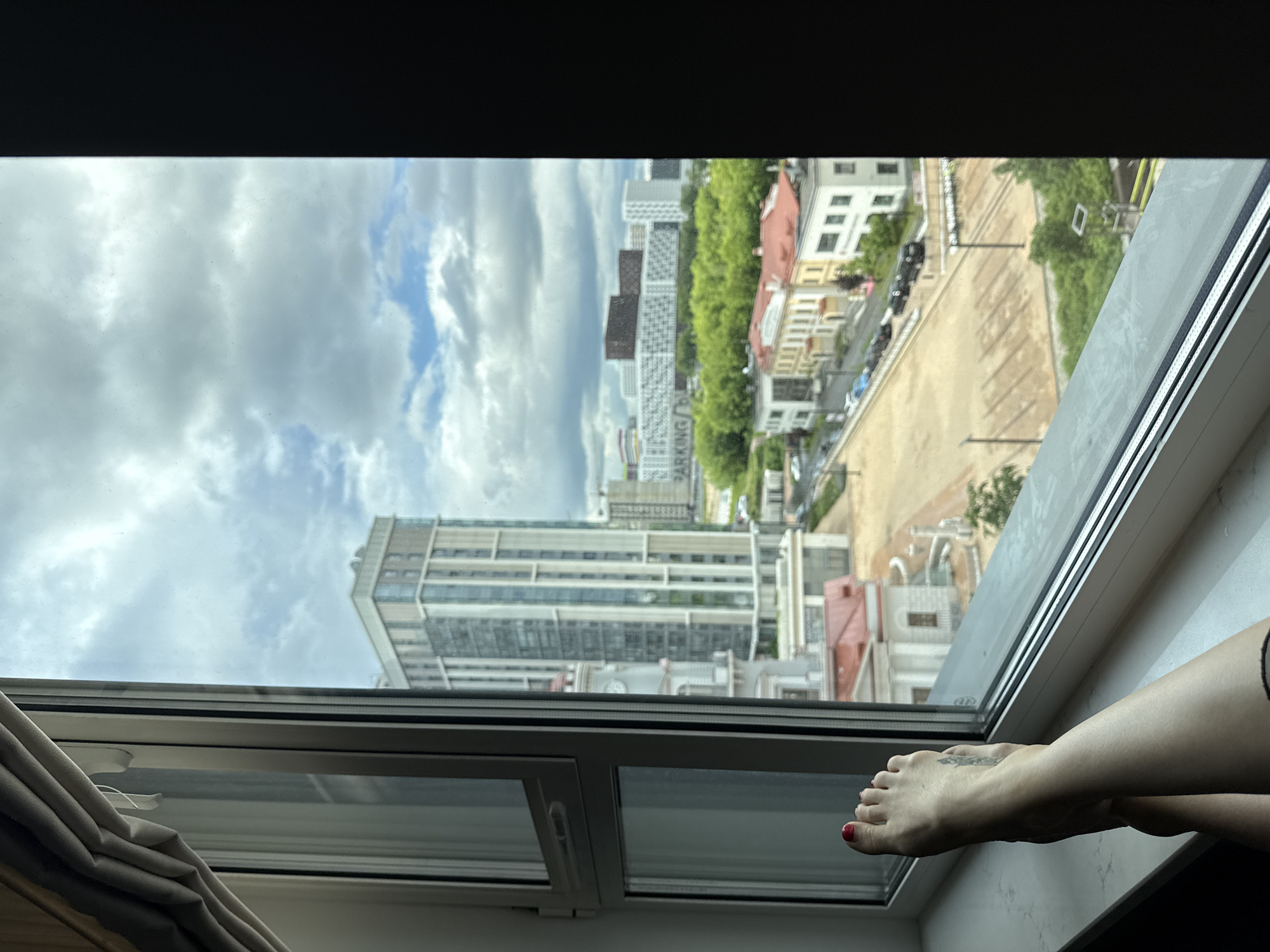 From an elevated perspective, a view outside a window reveals the city of Dolgoprudny, Russia. The architecture includes multi-story apartment buildings and older, smaller structures. A sky filled with clouds can be seen above the cityscape. A person's lower leg and foot, with red nail polish, are visible resting on the window frame. The lower part of the window frame and curtain can be seen.