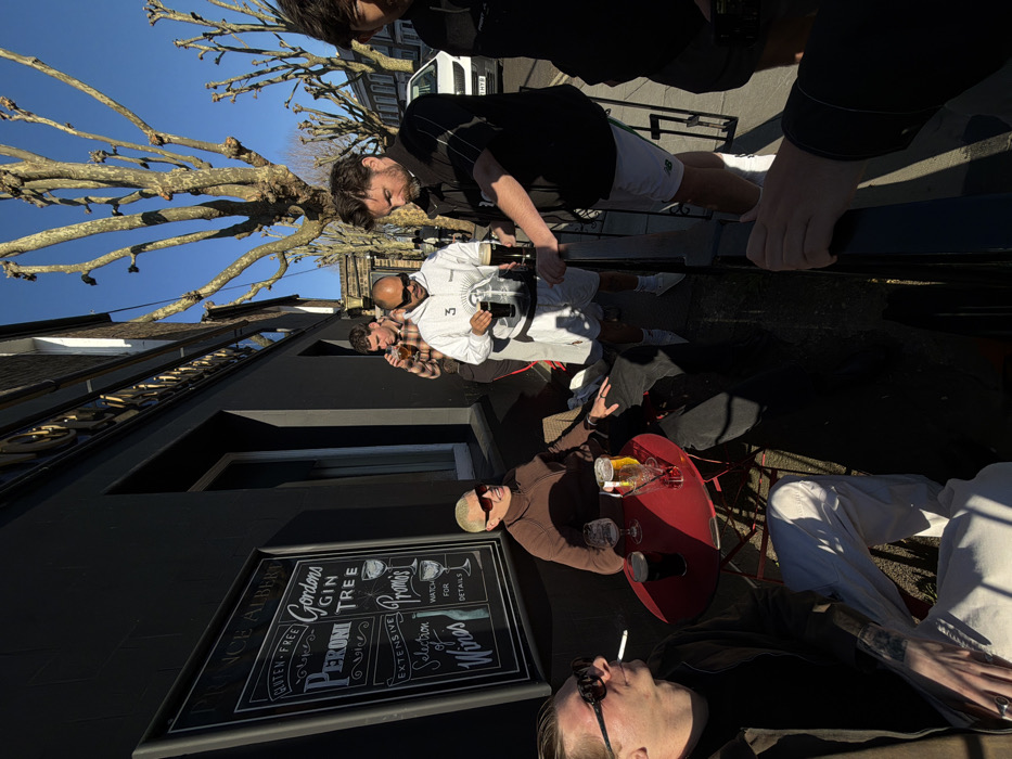 An outdoor scene depicts approximately six adult males socializing in front of a dark-colored building identified by a sign as "PRINCE ALBERT." The sign also advertises "Gluten-Free Peroni," "Gordons Gin Tree," "Extensive Selection of Wines," and "Promos." The location is a street in London, United Kingdom, characterized by bare deciduous trees and a clear blue sky, indicating a sunny day in a cooler season.
In the foreground, to the left, a person with blonde hair and sunglasses sits at a round red metal table, holding a dark beer glass and smiling. A lighter-colored beer and a clear glass are also on the table. Another individual, partially visible to the left, wears sunglasses and a dark jacket, holding a lit cigarette and looking away.
Towards the center-right, a man in a white hooded sweatshirt and white shorts wears sunglasses and holds a black tray with two dark beer glasses. He is in the process of handing one beer to another man dressed in a black polo shirt with white stripes and white shorts bearing a "NB" (New Balance) logo, who is reaching for the drink. Behind them, a man in a plaid shirt is seen drinking from a glass. Another man is partially visible on the far right. A white car is parked on the street in the background. A black railing separates the immediate foreground from the outdoor seating area.