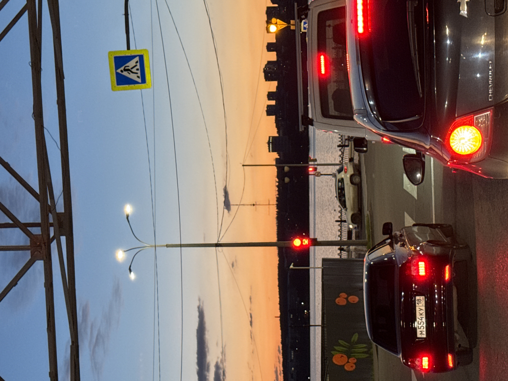 This is a medium shot, eye-level, vertical, outdoor, daylight, street-level photograph taken in Izhevsk, Russia. The image captures a traffic scene during twilight, likely on a bridge or embankment by a body of water, with a city skyline visible in the background.
In the foreground, a dark-colored sedan, possibly a Chevrolet based on visible badging, is stopped at an intersection, its brake lights illuminated. The car has a license plate that reads "M554KY 18". To the right of this car, another vehicle, a white van or SUV, is also stopped, with its brake light illuminated.
Further back, a beige sedan is seen approaching a pedestrian crossing. A traffic light on a pole is illuminated red. Above the traffic light, a pedestrian crossing sign is visible. Several overhead power lines crisscross the sky.
The background features a wide expanse of water, possibly the Izh River. On the opposite bank, a silhouetted cityscape with buildings of varying heights is visible. The sky above is a gradient of blues and oranges, indicative of sunset or twilight. Decorative elements, appearing to be painted orange spheres and green leaves on a fence, are visible on the left side of the image.
The overall impression is a calm, urban evening scene with traffic at a standstill. The lighting suggests it is late evening or early morning, with artificial lights from the city and vehicles beginning to dominate over the natural light.