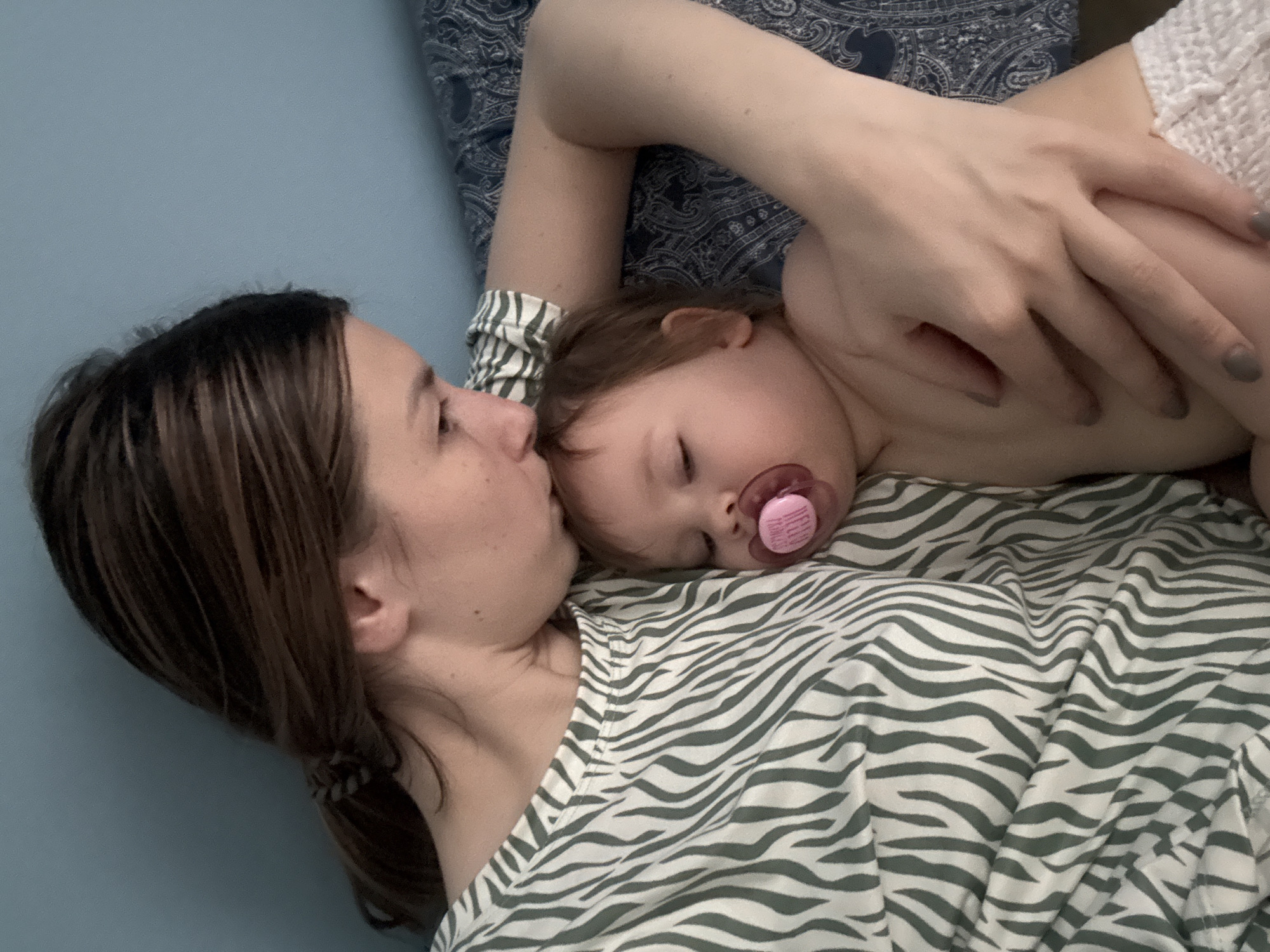 A woman lies on her back on a bed with zebra-striped bedding, with a young child asleep on her chest. The child has a pacifier in their mouth. The woman's right arm is draped over the child's body, her hand resting on the child's leg. The woman's hair is brown and pulled back in a ponytail. A blue wall is visible to the left of the woman's head. The location is not discernible from the image.