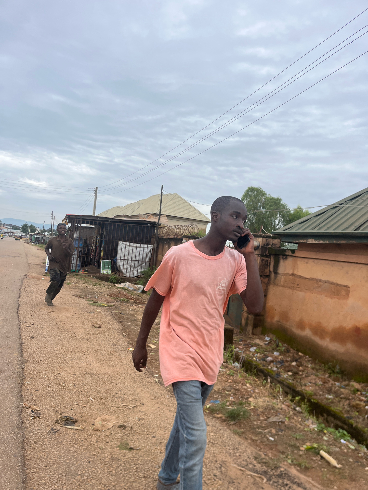 The image depicts an outdoor scene on a cloudy day in Barakin Akawo, Nigeria. A young man in a pink t-shirt and blue jeans is walking and talking on a mobile phone, holding it to his right ear. He is in the foreground and appears to be walking towards the viewer. To his left and further down the dusty, unpaved road, another man in a dark t-shirt and pants is running, looking towards the camera with a smile. The environment consists of a road with a gravel shoulder, lined with buildings, some with corrugated metal roofs and barred windows. There is debris and vegetation along the side of the road. Power lines are visible against the sky.