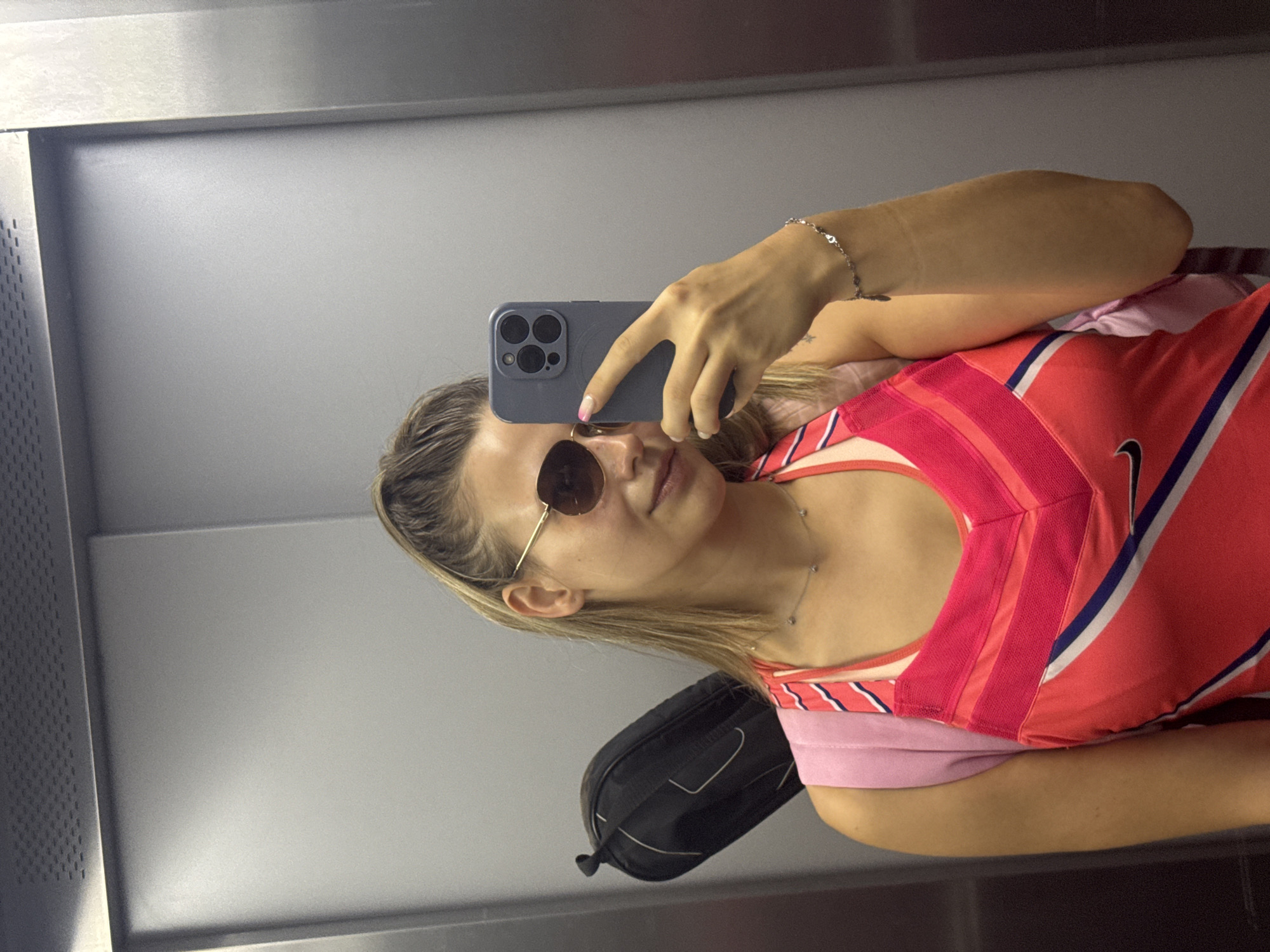 The image depicts a young woman taking a selfie in an elevator. She is wearing a pink and red athletic top, sunglasses, and a silver necklace. A pink backpack is visible on her shoulders, with a black bag resting behind it. The elevator interior is metallic and gray. The scene is likely a public space within Sofia, Bulgaria, possibly a hotel or residential building elevator, given the typical urban setting of Sofia. There are no other people or any distinctive urban landmarks present in the image.