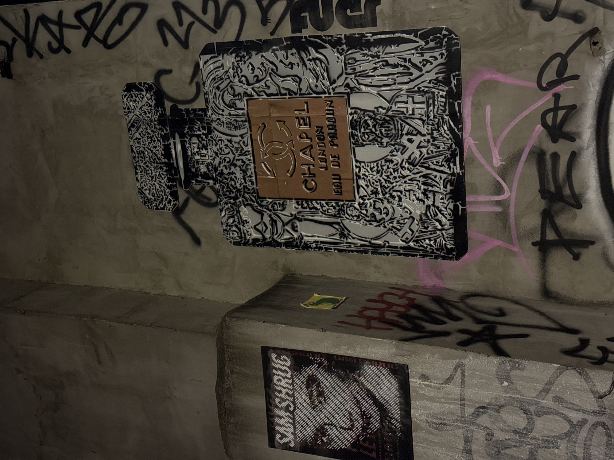 The image shows a concrete wall in what appears to be an urban environment, possibly an alleyway. The wall is covered in graffiti, including abstract markings and lettering. A large, black-and-white stylized representation of a perfume bottle, labeled "Chapel London Eau de Parduin," is prominently displayed. Below this, on a ledge, is a poster featuring a pixelated portrait with the name "Sam Shrug" printed alongside it. A small yellow rectangle (possibly a sticker or piece of paper) is also visible on the ledge. The overall atmosphere is gritty and urban, consistent with street art in a city setting. There are no people visible in the frame. There is not enough information in the image to confirm that the location is London, United Kingdom.