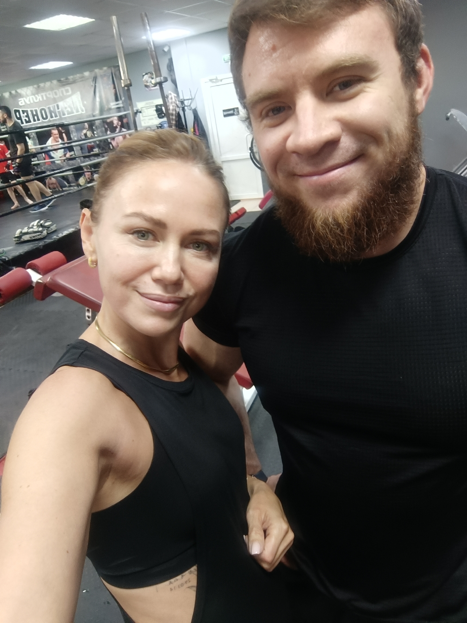 The image shows a selfie of a light-skinned woman and a light-skinned man inside what appears to be a gym. The woman is on the left side of the frame and is wearing a black athletic top. She has fair skin and her hair is pulled back. The man is on the right side of the frame and is wearing a black t-shirt. He has a beard and lighter colored hair. In the background, a boxing ring is visible with people inside. Gym equipment can also be seen. The photograph was likely taken at the "Медведь" Fighting Club in Naberezhnye Chelny, Russia, as this is suggested by the background, including the boxing ring and signage.