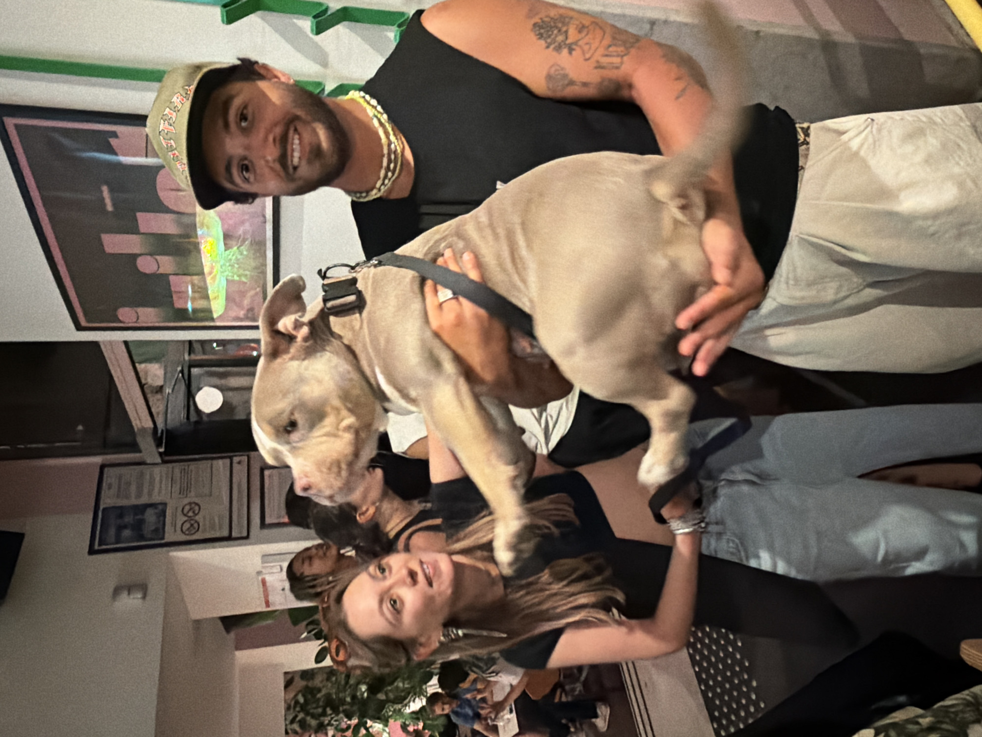 A man and a woman are posing for a photograph with a dog inside a building. The man, on the right, is wearing a black sleeveless shirt, a green cap, and multiple necklaces. He is holding a light-colored dog, which is looking to the left. The woman, on the left, is wearing a black top and light blue jeans. She is looking at the dog and appears to be smiling. In the background, other people are visible, as are posters on the wall and some plants. The environment suggests an indoor public space, possibly a bar or a restaurant. No specific details in the image indicate the city of Lyon, France.