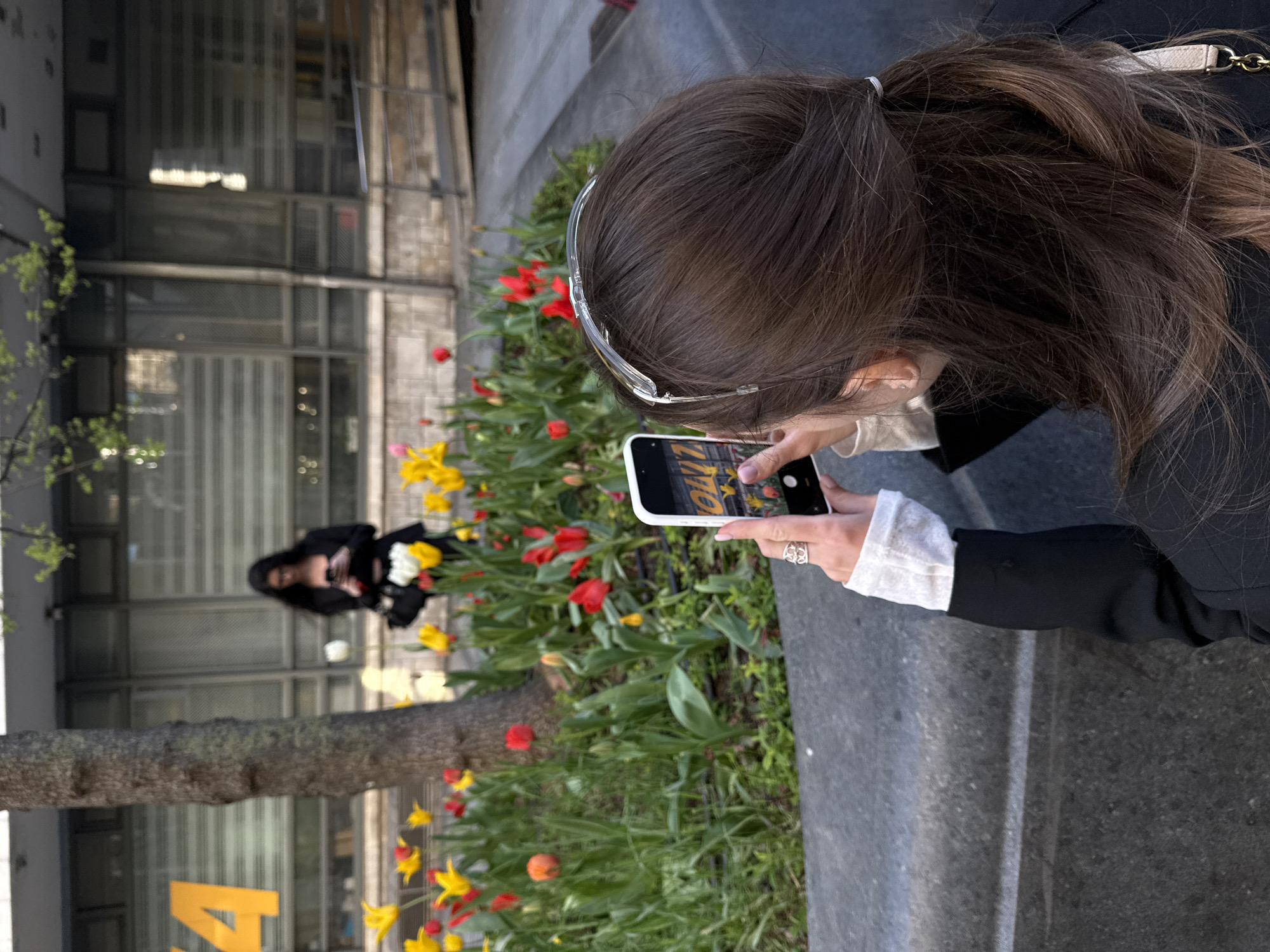 A woman is taking a picture with her smartphone of another woman posing in front of a flower bed filled with tulips. The photographer is wearing clear glasses and a dark jacket with white lace cuffs. The person being photographed is wearing a dark coat and holding a bouquet of flowers. The flower bed contains red, yellow, and orange tulips. In the background, there is a building with a grid-like facade and a tree with new leaves growing on it. A yellow sign is visible on the side of the building. The location is likely urban. This is not in Belgrade, Serbia.