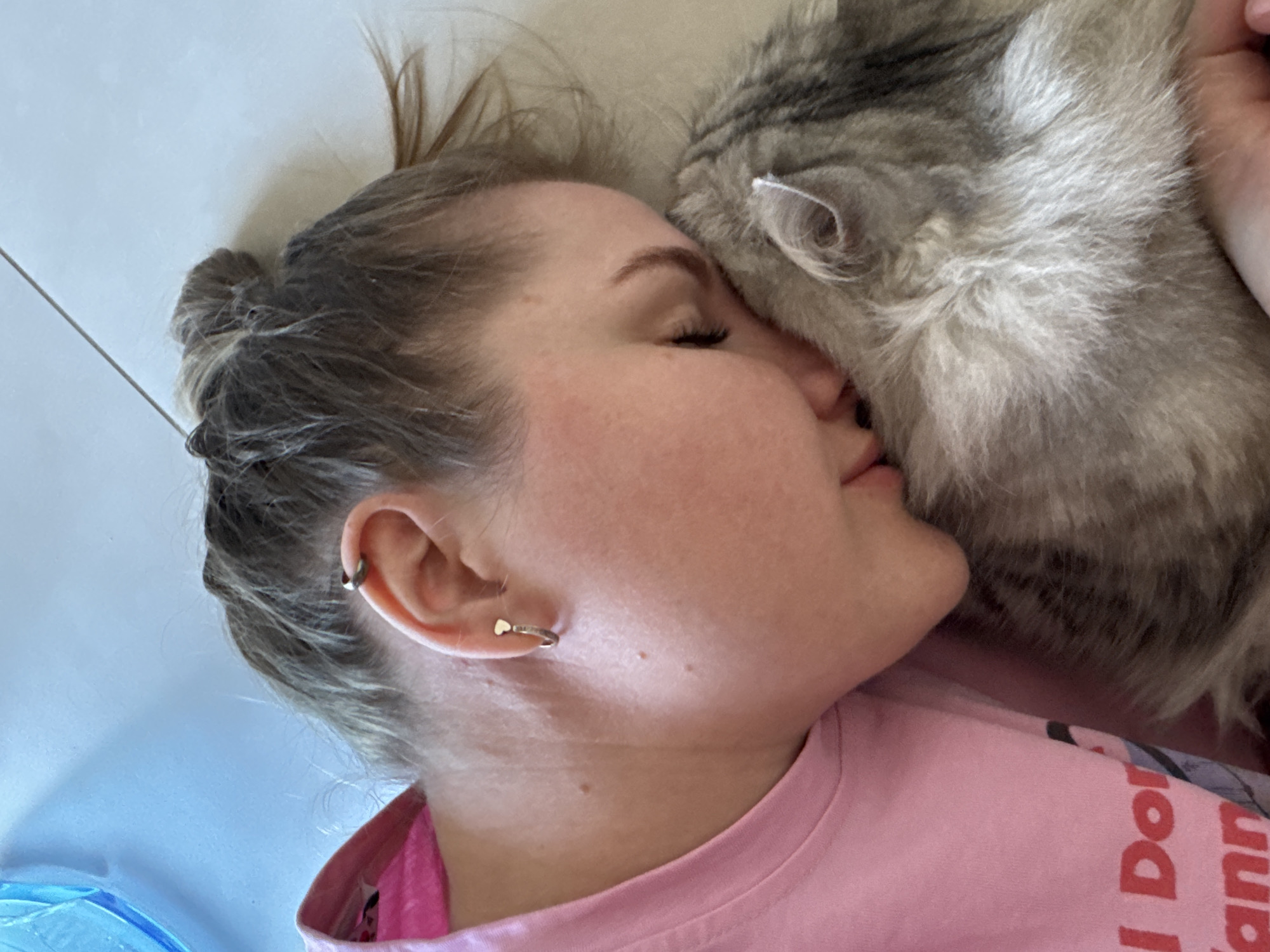 An individual is depicted in an indoor environment in Gornyi (Dvurechenskoe S/P), Russia. The subject, a human female, is positioned on her right side, with her face pressed against a domestic feline. She is wearing a pink t-shirt with red lettering partially visible, reading "I Don" and "ann". Her medium brown hair is styled in a bun, with some loose strands. The visible right ear features two piercings: a silver helix piercing with a heart-shaped stud, and a silver hoop in the lobe. Her eyes are closed or mostly closed. The feline subject is a long-haired cat, primarily grey and white, with its head positioned against the human's face. The immediate background consists of a light-colored, possibly tiled floor, with a visible grout line. In the upper right quadrant, a translucent blue object is partially visible. The scene captures a close physical interaction between the human and the cat.