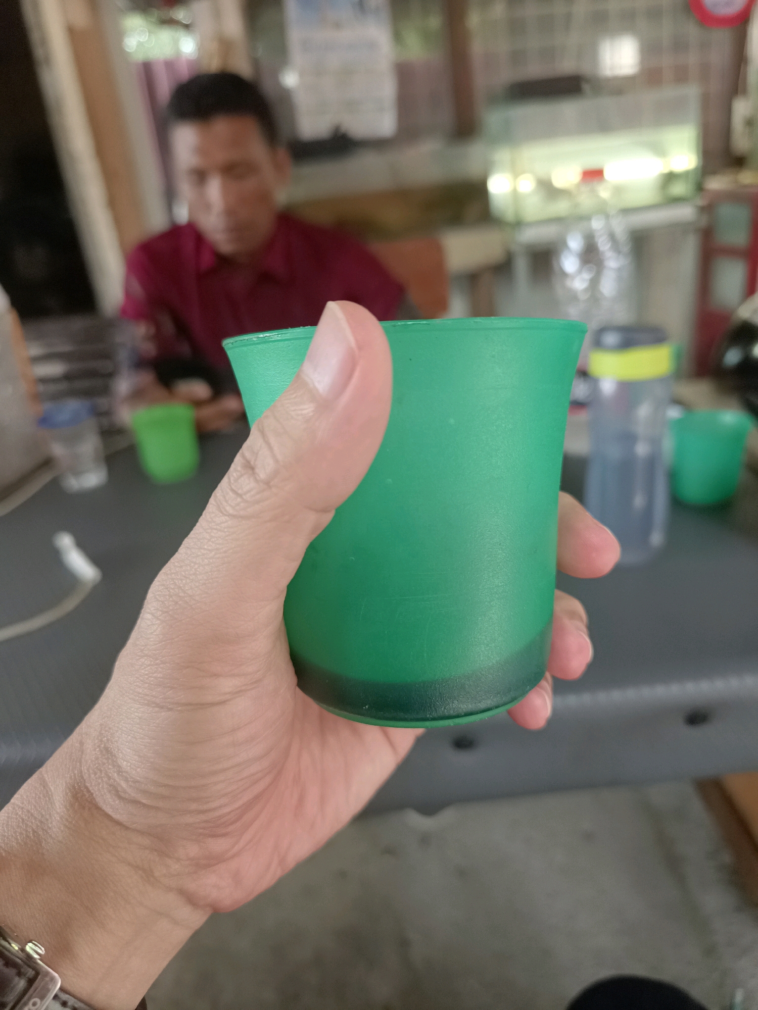 A close-up shot features a person's hand holding a small, opaque green plastic cup. The cup contains a dark liquid, filling the bottom quarter of its volume. The hand is positioned in the foreground, with the fingers curled around the cup. In the background, out of focus, a man wearing a maroon shirt is seated. The environment appears to be an indoor or semi-outdoor setting, possibly a casual eatery or gathering space, with other objects such as bottles and cups visible on a table. The image was taken in Mesjid Reuntoh, Indonesia.