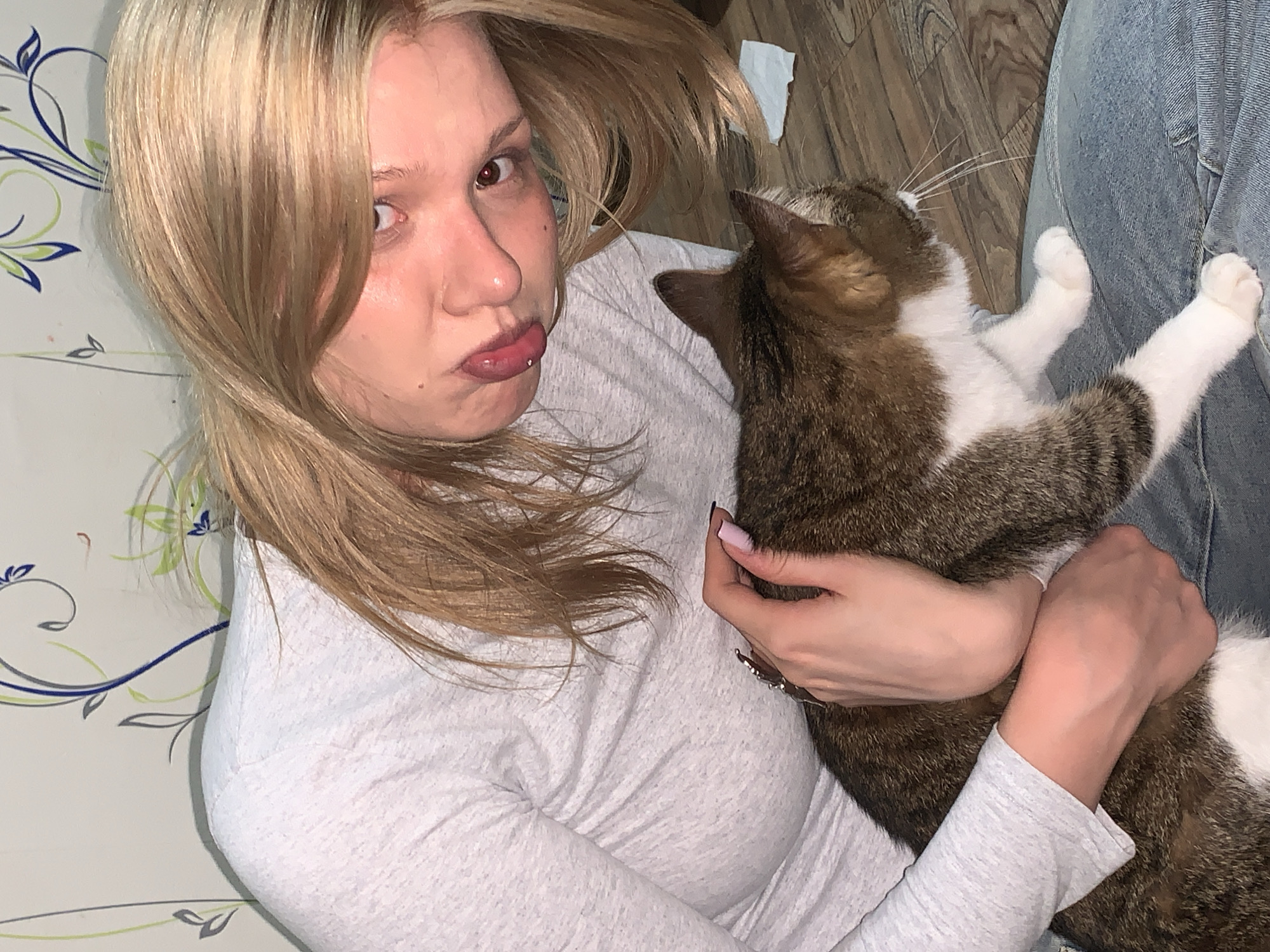 A blonde woman with a lip piercing lies on a patterned surface (possibly a couch) in what appears to be an indoor environment, holding a brown and white cat. The cat has its paws extended. The woman is looking at the camera with a slightly pouty expression. A person wearing denim clothing is visible partially in the frame. The location is Chaikovsky, Russia.