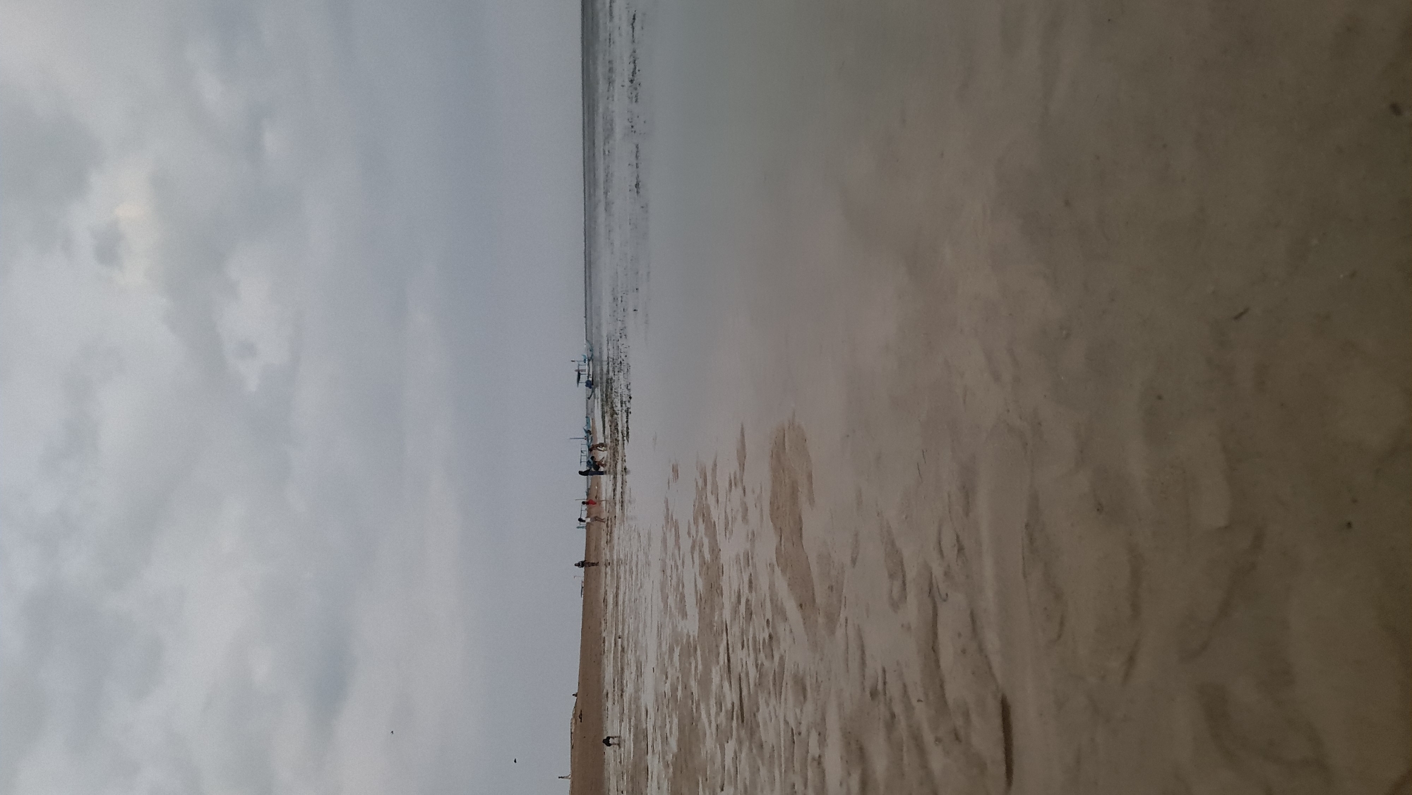 The image depicts a low-tide beach scene, likely in Denpasar, Indonesia, under an overcast sky. The foreground is dominated by wet sand with visible ripples and patterns left by receding water. In the middle ground, a group of people, including adults and children, are scattered along the sandy shore and near the water's edge. Some appear to be walking, while others are interacting or observing the surroundings. Several traditional Balinese jukung boats, characterized by their outriggers, are visible anchored in the shallow water or near the shoreline. The water is calm and reflects the muted light of the cloudy sky, with areas of darker, vegetated seabed showing through. The overall atmosphere is serene and suggests a typical coastal activity during a cloudy day.
