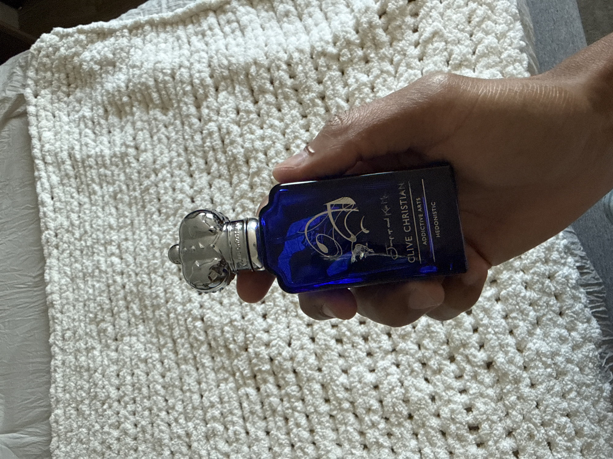 A hand holds a Clive Christian perfume bottle against a background of a textured, off-white blanket. The bottle is dark blue with silver accents and lettering. The blanket has a repeating pattern, possibly knitted or crocheted. The location is indeterminate, and there is no information to suggest it is in Palm Springs, United States of America.