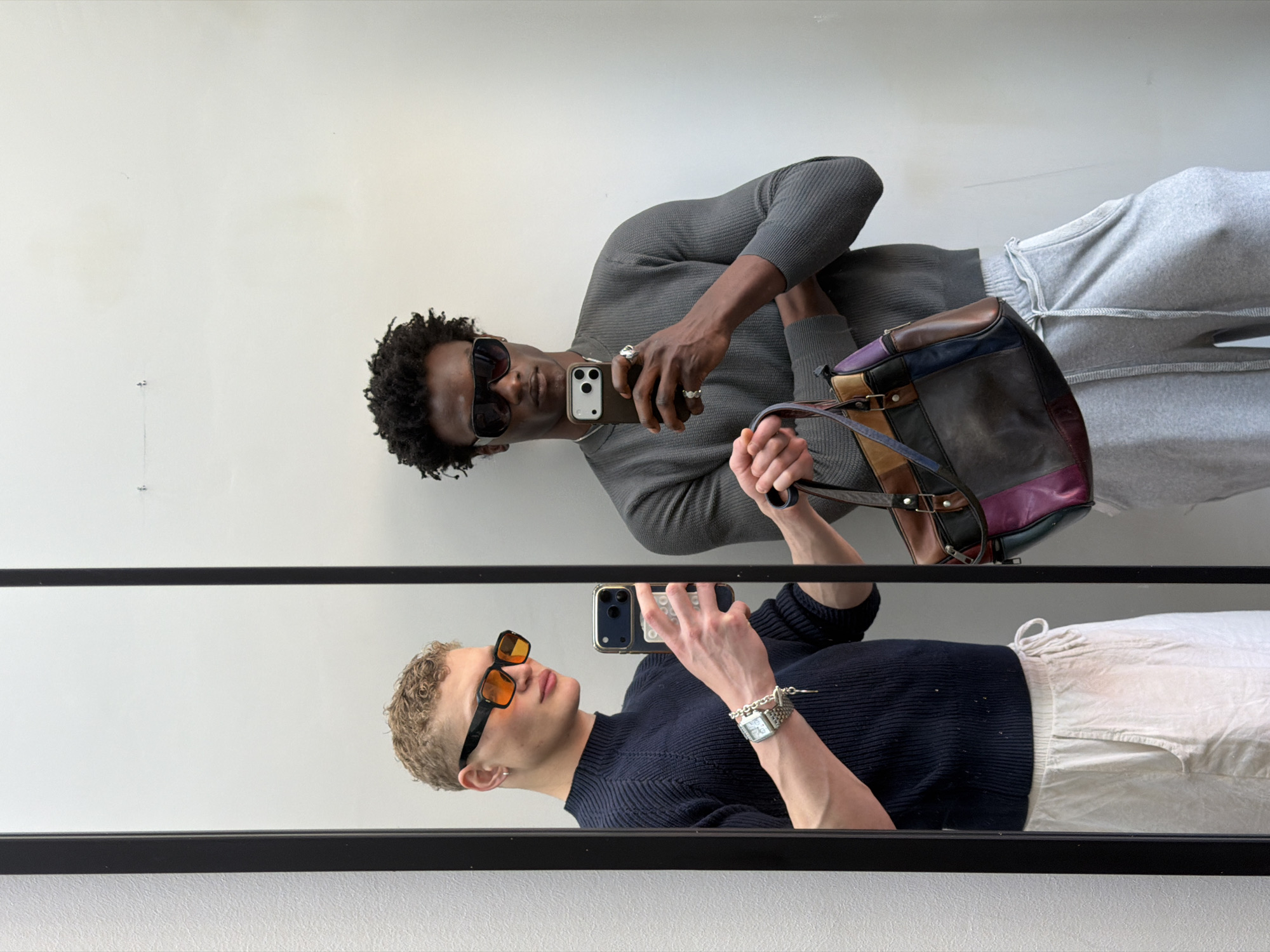 A man with dark skin, wearing sunglasses, a gray sweater, and gray sweatpants, holds a smartphone in his left hand and a multicolored leather bag in his right, facing a mirror. Reflected in the mirror is a second man with light skin, wearing orange-tinted sunglasses, a navy sweater, and white pants, also holding a smartphone. The mirror has a black frame. The location is most likely a studio setting in London, United Kingdom, judging by the neutral background and style. Both men appear to be taking selfies in the mirror.