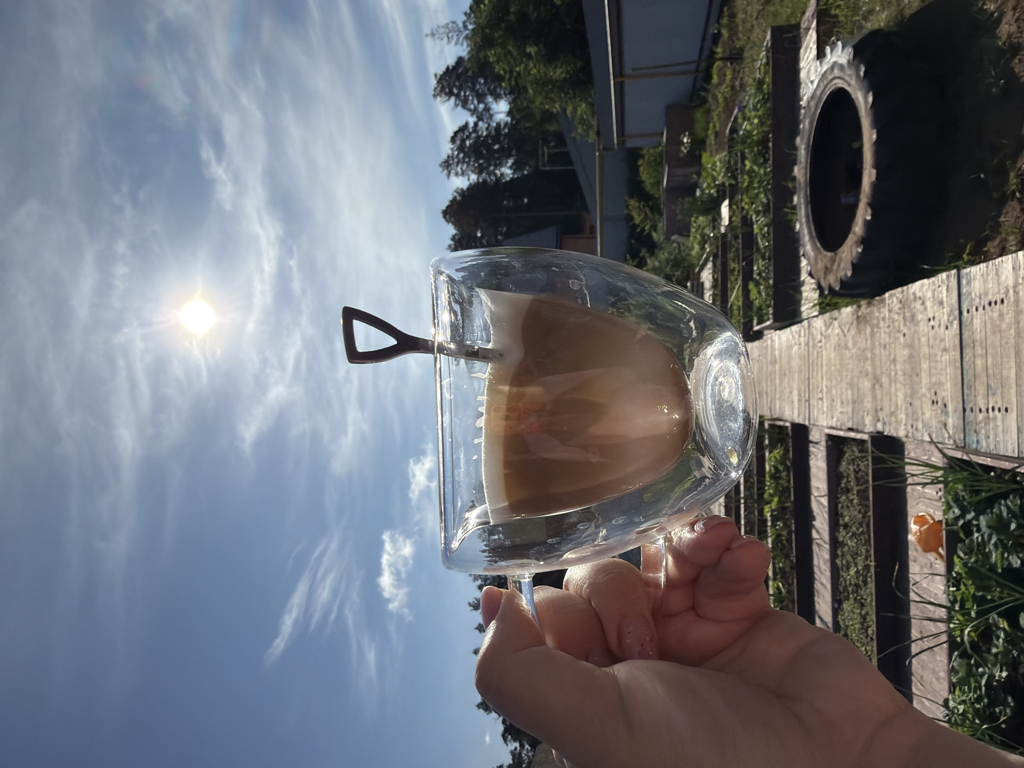 A hand holds a glass cup filled with a beverage against a blue sky with scattered clouds and the sun. The glass contains a light brown liquid, possibly coffee with milk. A spoon rests inside the cup. In the background, there is greenery, trees, and a fence. To the right, a tire is visible. The location is possibly Chaikovsky, Russia, based on external knowledge of similar outdoor settings in that region. The image depicts a leisurely moment of enjoying a drink outdoors.