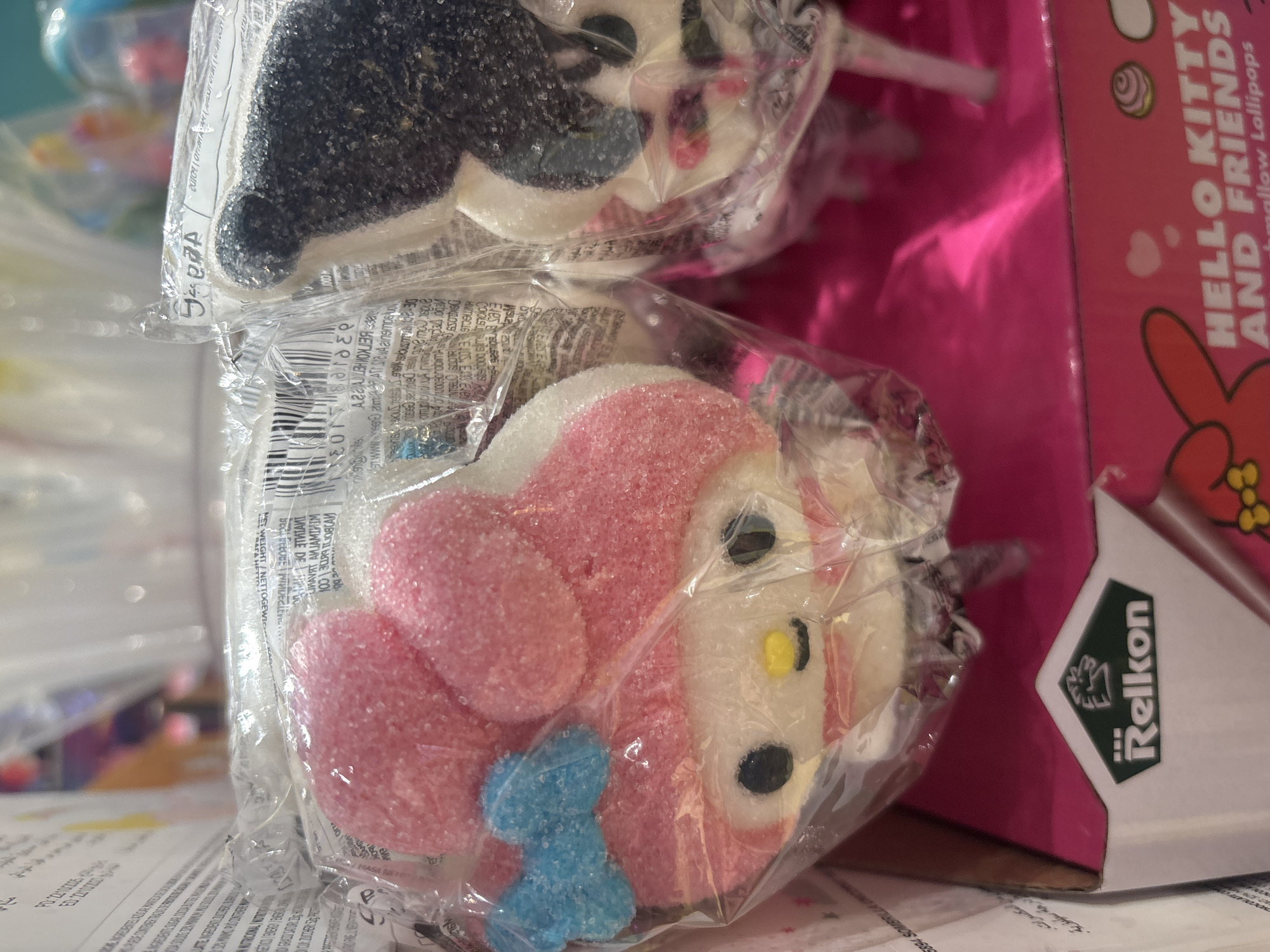 Two individually wrapped marshmallow lollipops shaped like cartoon characters are displayed on a pink box in a retail environment. The lollipop on the left features a pink character with a blue bow and two pink ears. The lollipop on the right has a black and white character with black ears. Both lollipops are covered in sugar crystals and are made of marshmallow. Text on the pink box includes "Reikon" and "HELLO KITTY AND FRIENDS Marshmallow Lollipops". The background shows shelves with more packaged candy. This scene is likely in Dubai, United Arab Emirates.