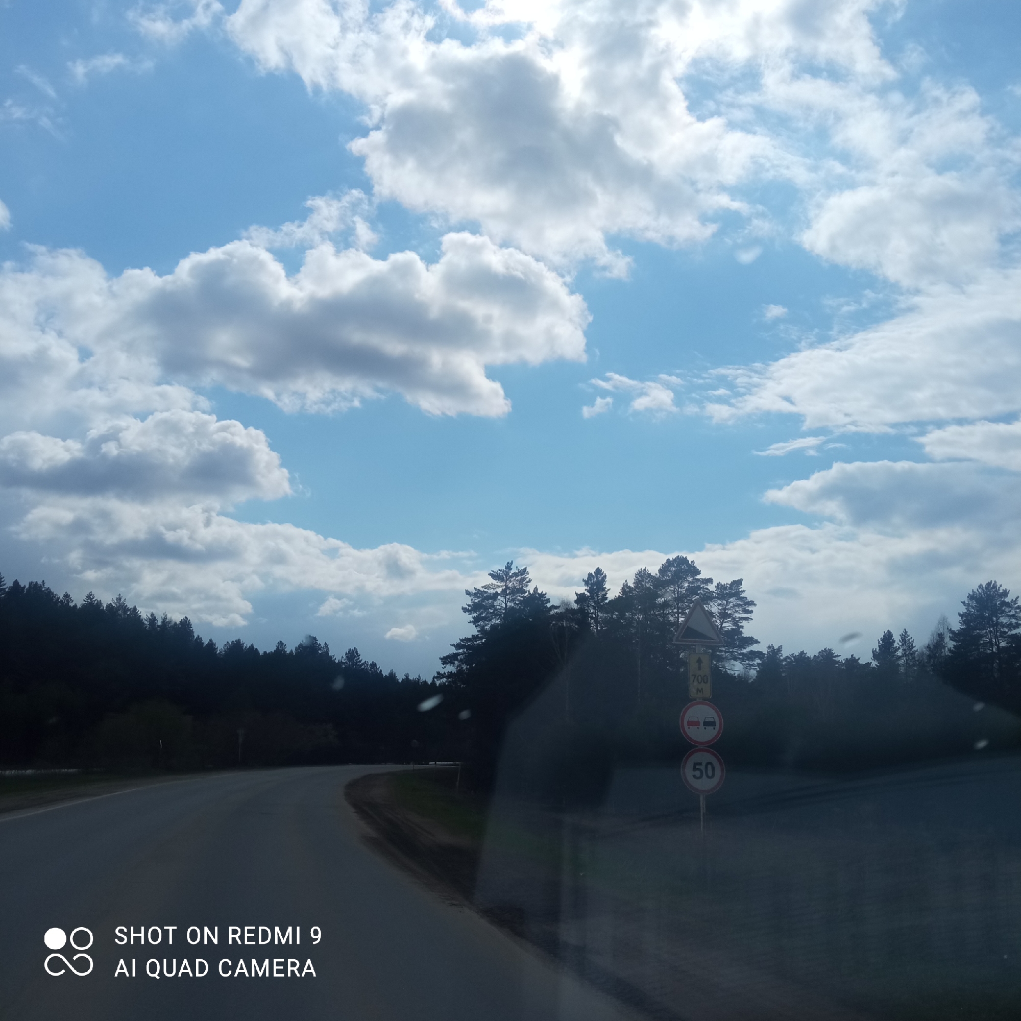 The image presents a daytime view from inside a vehicle on a rural road in Прикамский, Russia. The sky above is largely covered by large, puffy white and grey cumulus clouds, with bright patches of blue visible between them, suggesting mixed weather conditions.
A dense, dark green forest of coniferous trees lines both sides of the asphalt road, extending towards the horizon. The road itself is dark grey and appears to curve slightly to the left in the foreground. There are indications of wetness or reflections on the surface through which the photo was taken, particularly in the lower right quadrant.
On the right side of the road, a pole holds several traffic signs. From top to bottom, these include a yellow rectangular sign with an upward arrow and "700 M," likely indicating an upcoming feature 700 meters ahead. Below it is a circular sign with a red border depicting two cars, signifying "No Overtaking." The lowest sign is a circular speed limit sign with a red border and the number "50," indicating a maximum speed of 50 km/h.
In the bottom-left corner, a watermark reads "SHOT ON REDMI 9 AI QUAD CAMERA." The scene depicts a natural landscape with clear roadway infrastructure, captured under a bright, dynamic sky.