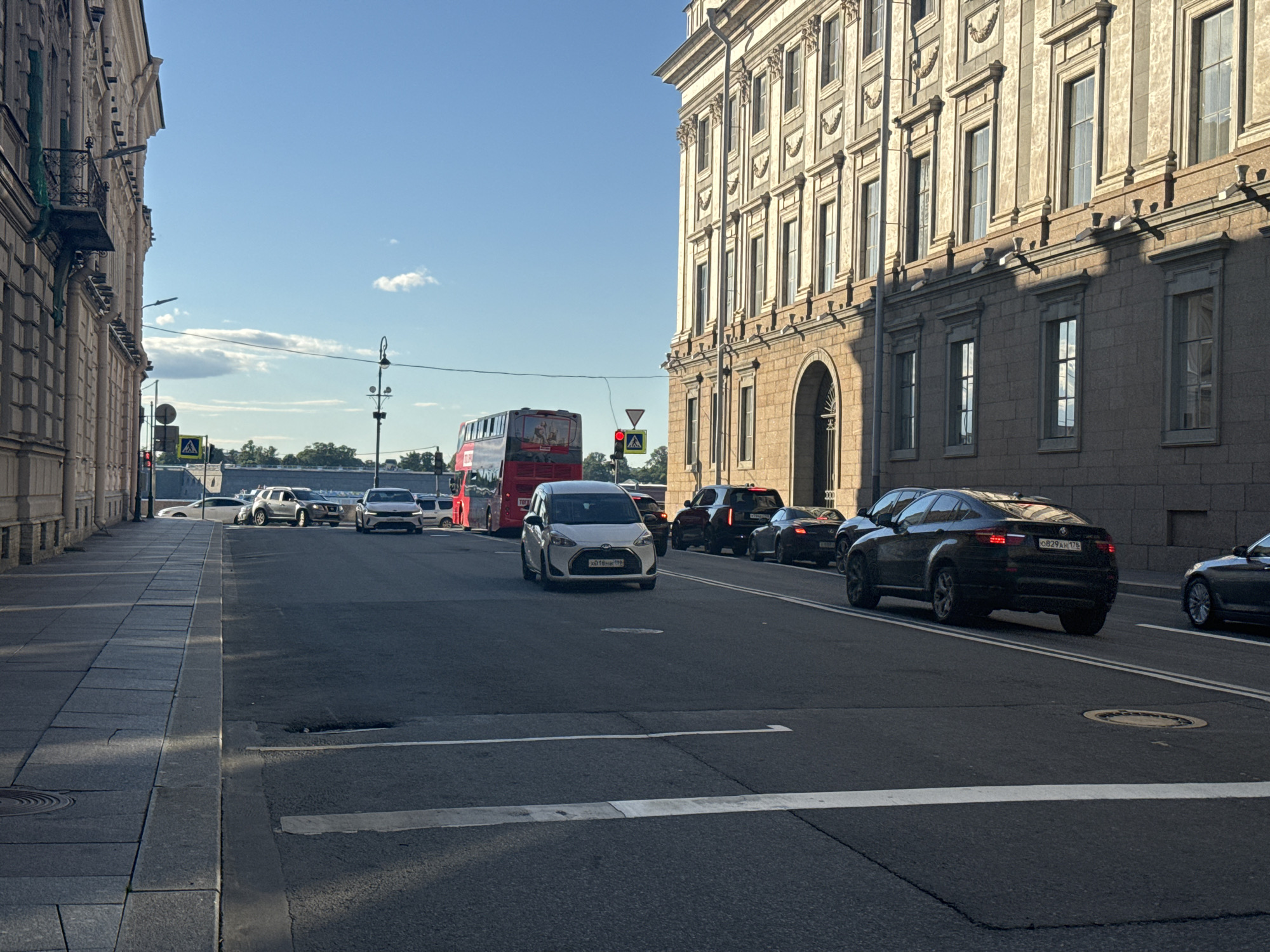 The image depicts a street scene in Saint Petersburg, Russia, likely along Nevsky Prospekt, given the grand architecture and the presence of a double-decker bus. The photograph is taken from street level, looking down a wide avenue lined with impressive, ornate buildings on the right and a sidewalk with more subdued architecture on the left.
In the foreground, the asphalt road is visible, marked with white lines indicating lanes and a pedestrian crossing. The left side of the image features a grey stone sidewalk with rectangular paving stones. Sunlight casts long shadows across the road and sidewalk, suggesting it is late afternoon or early morning.
Several vehicles are present on the road. A white hatchback is in the center lane, facing the viewer. To its right, a black SUV is moving away from the camera. Further down the road, a red double-decker bus is also visible, suggesting it is a tourist bus. Other vehicles, including a silver SUV and a white sedan, are parked or moving in the distance. The architectural style of the buildings, characterized by large windows, intricate detailing, and a sense of historical grandeur, is typical of Saint Petersburg. The sky above is a clear blue with a few wispy clouds.