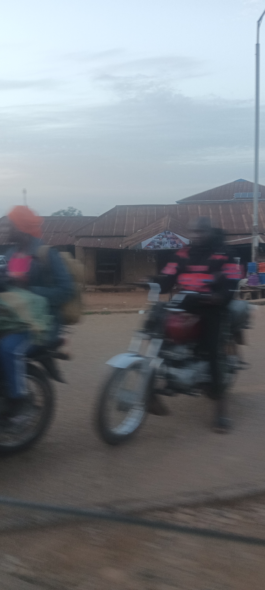 Two motorcycles in motion, ridden by individuals, are depicted in the foreground. In the background is a street scene, featuring structures with corrugated iron roofs, indicative of commercial establishments. A utility pole is visible. The setting is likely a street in Mangu Town, Nigeria.