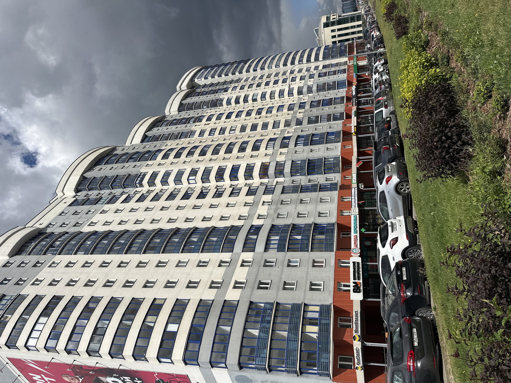 The image shows a modern, multi-story residential building with a curved facade, located in Minsk, Belarus. The building is predominantly white and gray with blue accents on the windows and balconies. The ground floor features a row of storefronts with signs in Cyrillic script, indicating businesses such as a bank and a pharmacy. A line of parked cars is visible in front of the building, and a grassy median with bushes separates the road from the building's entrance. The sky above is partly cloudy, with dark storm clouds gathering. The overall impression is of a contemporary urban landscape under an approaching storm.