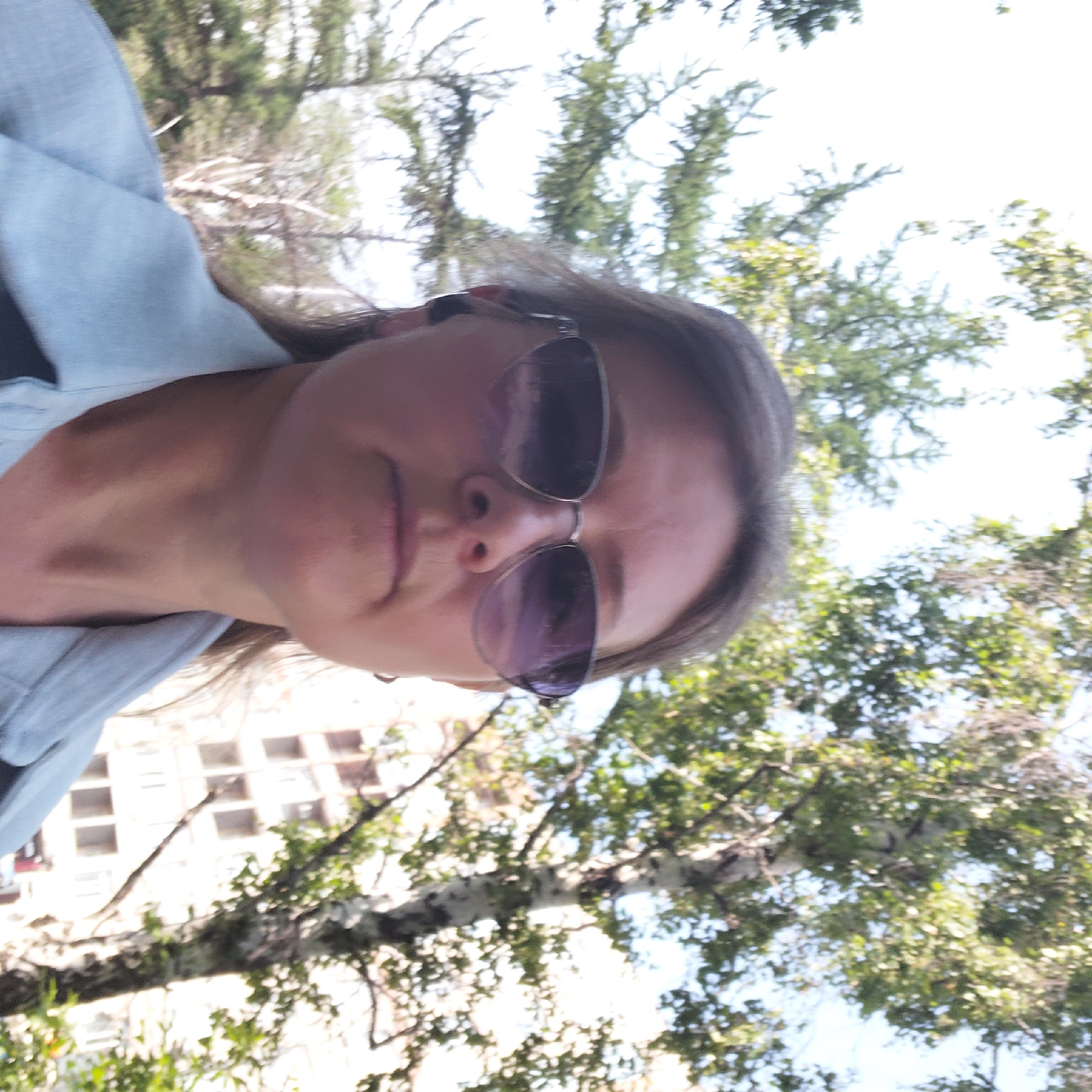 In this low-angle, close-up photograph, a woman with medium-length brown hair and sunglasses is captured from the chest up. She is wearing a light blue collared shirt. The background features a blurred view of trees and a building. The overall scene suggests an outdoor urban environment in Novosibirsk, Russia. The lighting indicates it is daytime.