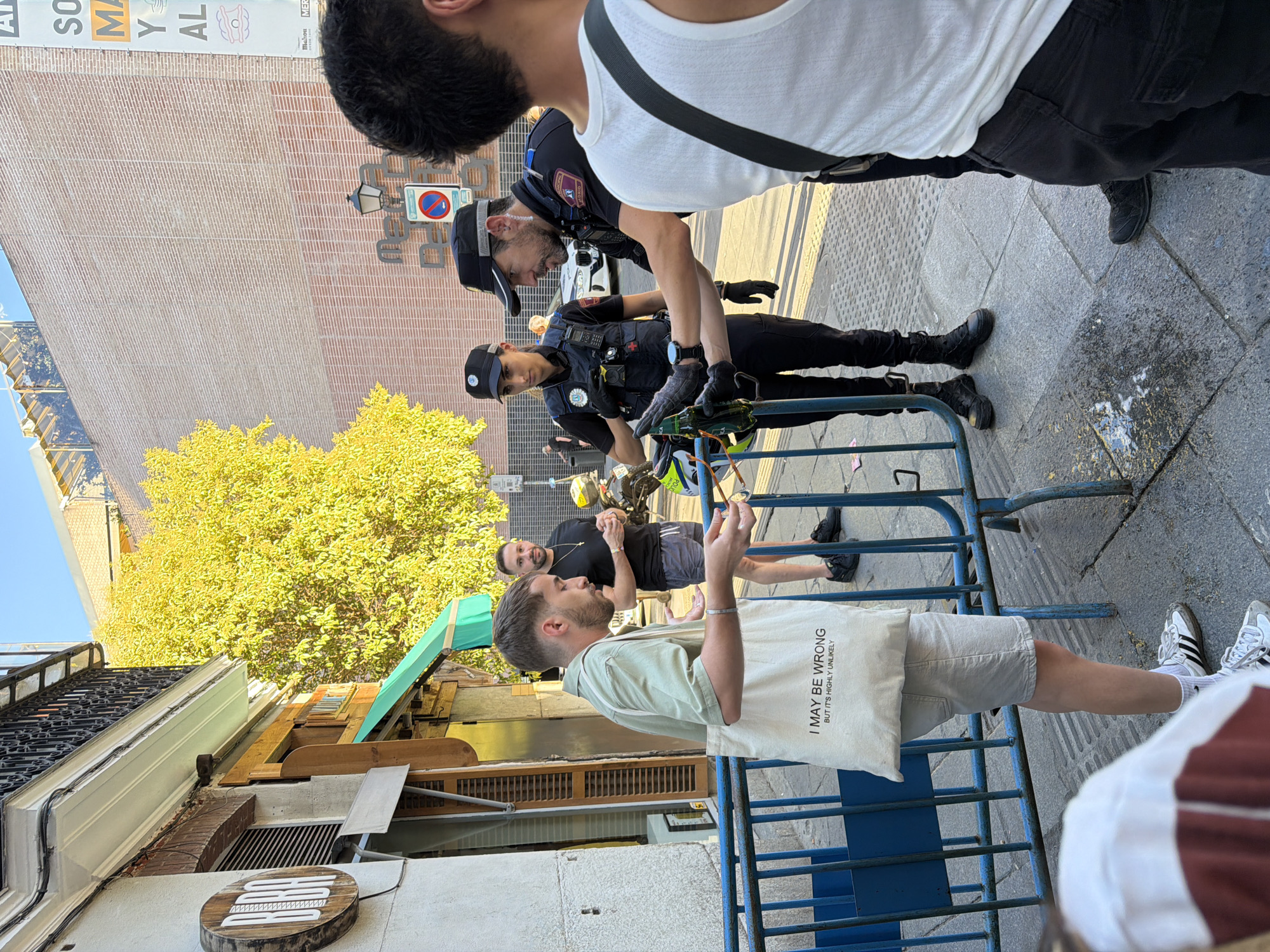 A man in a light green shirt and white tote bag is speaking with two police officers on a city street in Madrid, Spain. The man is wearing a tote bag with the text "I MAY BE WRONG BUT IT'S HIGHLY UNLIKELY" printed on it. One of the police officers, a woman, is standing to the left of the man, wearing a dark uniform with a badge and utility belt. The other police officer, a man, is to the right of the woman, also in uniform, and is handing a bottle to the man in the green shirt. Both officers are wearing black baseball caps and dark pants. In the background, there is a building with a yellow tree in front of it, and a blue metal barrier. A white car is visible parked behind the officers. The ground is paved with cobblestones.