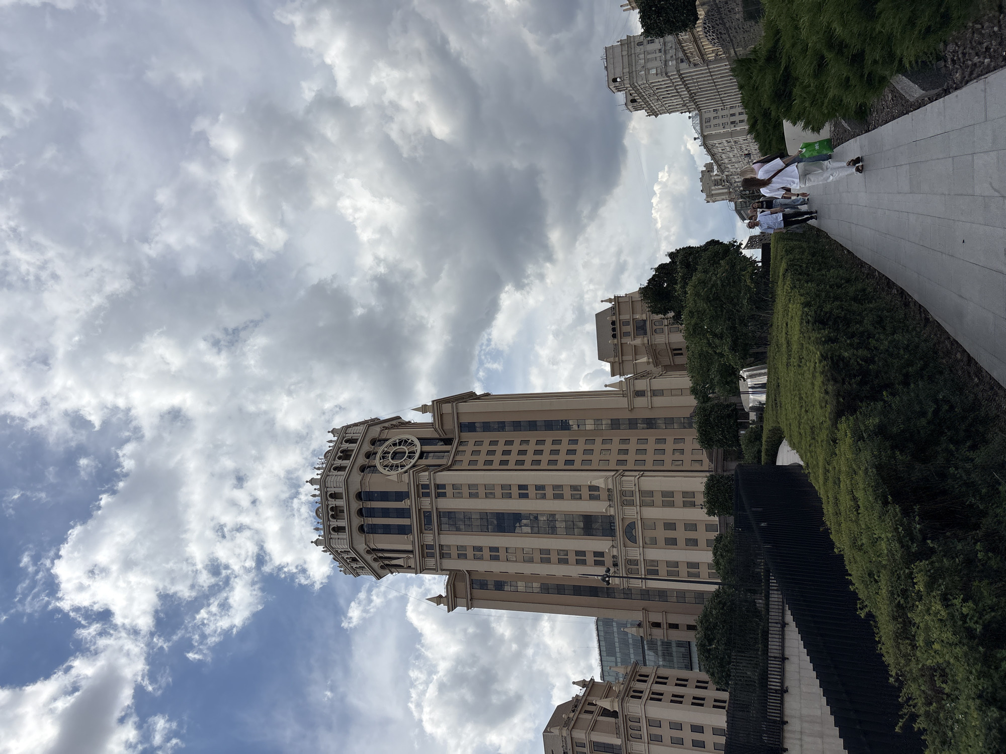 The image depicts a scene in Moscow, Russia, featuring the iconic Kotelnicheskaya Embankment Building, a Stalinist skyscraper, in the background. The building, characterized by its distinctive spire and ornate architecture, dominates the skyline under a cloudy sky. In the foreground, a paved walkway curves through manicured green bushes and hedges. Several individuals are walking along the path. One person, wearing a white t-shirt and white pants, carries a green shopping bag. Other figures are walking ahead, and a few more individuals are visible in the distance. The environment suggests a public park or promenade area adjacent to the building.