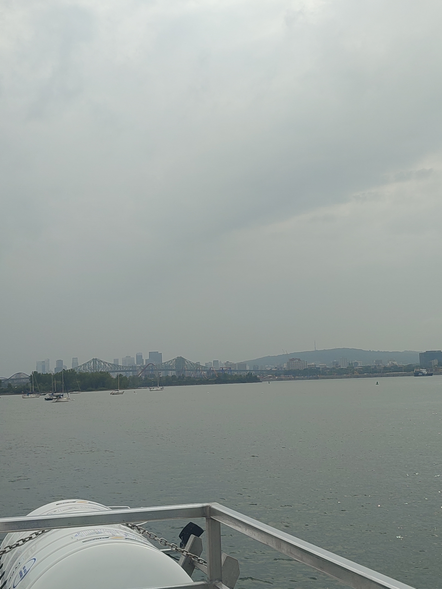 This media file captures a hazy, overcast day scene from the perspective of a boat. In the foreground, the top of a white, cylindrical object, possibly a life raft, is visible, secured by a chain. Beyond this, a calm, gray body of water stretches towards a distant cityscape.
The cityscape is dominated by several bridges, including what appears to be the Jacques-Cartier Bridge and the Victoria Bridge, spanning the water. A line of buildings and trees forms the shoreline, with more distant hills and structures suggesting a sprawling urban area. Several sailboats are scattered across the water, their masts tall against the muted sky. The weather appears to be cloudy and possibly humid, contributing to the low visibility. The time of day is indeterminate due to the overcast conditions. The scene is one of peaceful, albeit slightly somber, maritime activity. There is no discernible text in the image. The location context is Longueuil, Canada, which is consistent with the visible bridges and urban landscape.