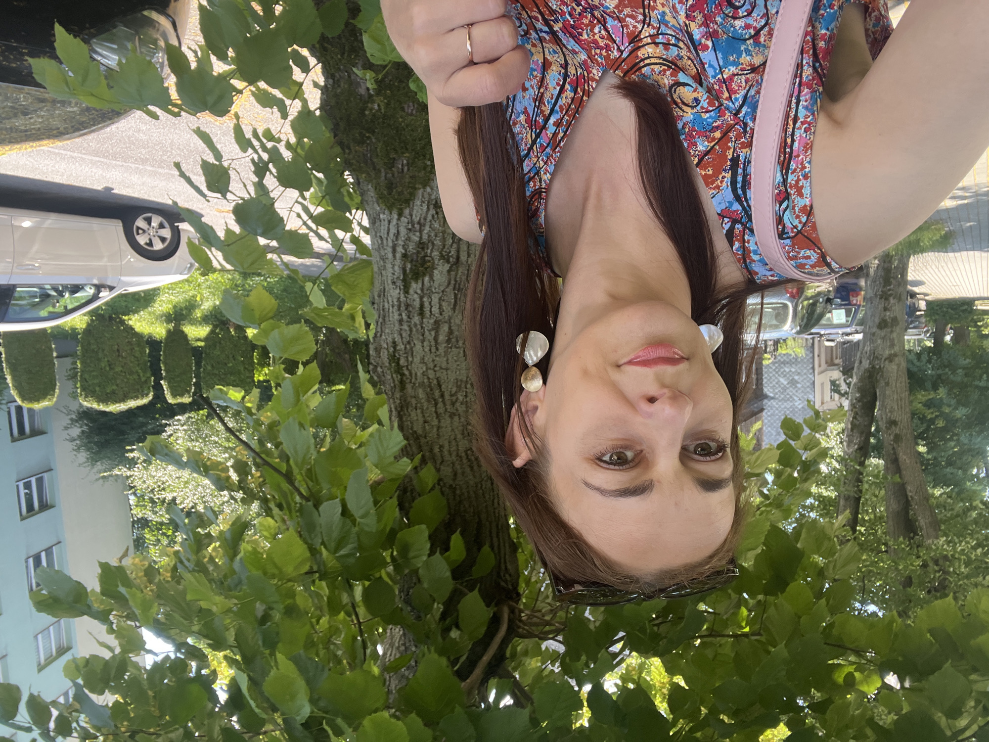 A woman is taking a selfie outdoors. She has long brown hair and is wearing a colorful patterned dress and large earrings. She is also wearing a ring on her left hand. Behind her, there are trees with green leaves, a tree trunk, and a building with windows. The image appears to have been taken during the daytime. The woman's face is tilted upwards towards the camera, and her expression is neutral. The scene appears to be a park or a street with greenery.