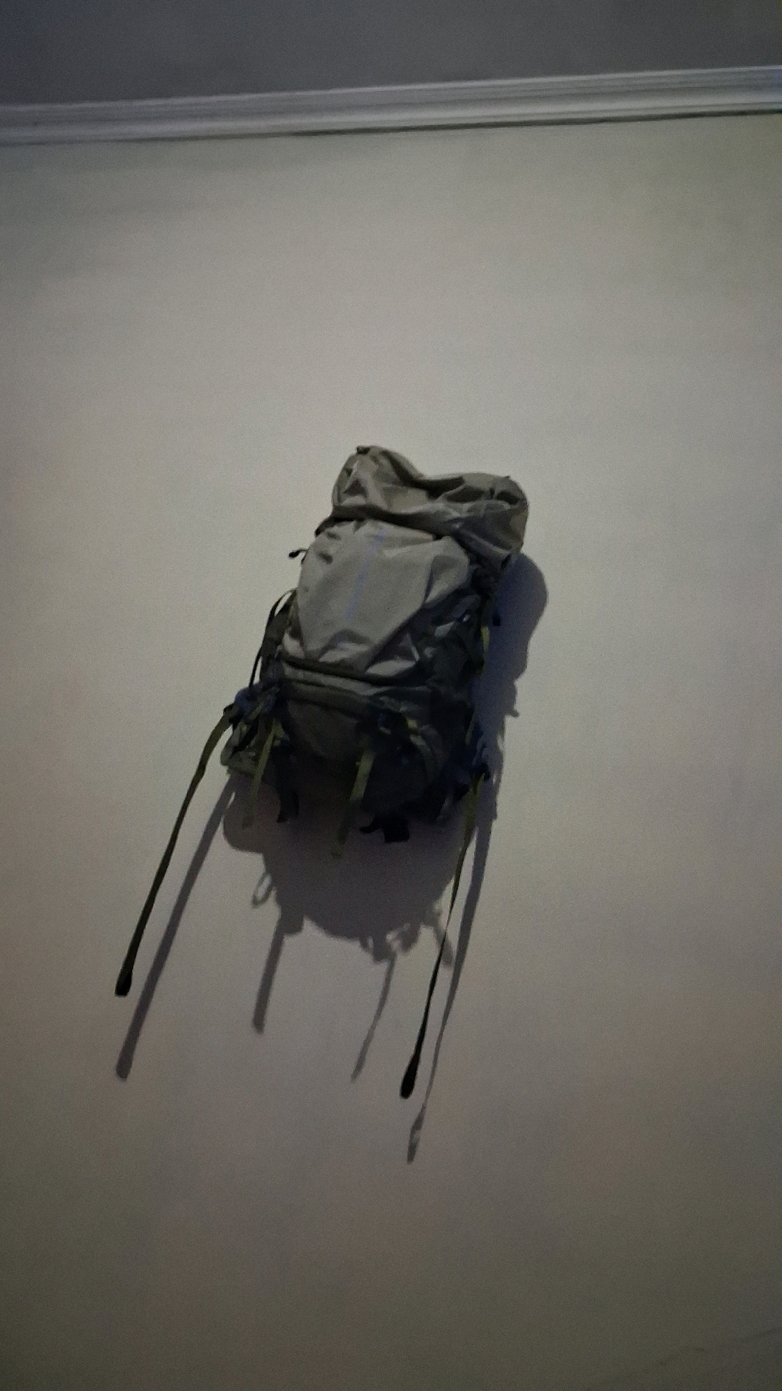 A grey backpack with green straps hangs from a wall in an interior space. The backpack is positioned towards the left side of the frame, casting a shadow to its right. The wall appears to be a light color and there is a subtle white molding along the top of the wall, suggesting a room in a building, possibly a residence or a storage area. The lighting conditions create a strong contrast between the backpack and its shadow, making the shadow prominent on the wall. The image does not provide any information about the city of Babakan, Indonesia, or any specific location within it.
