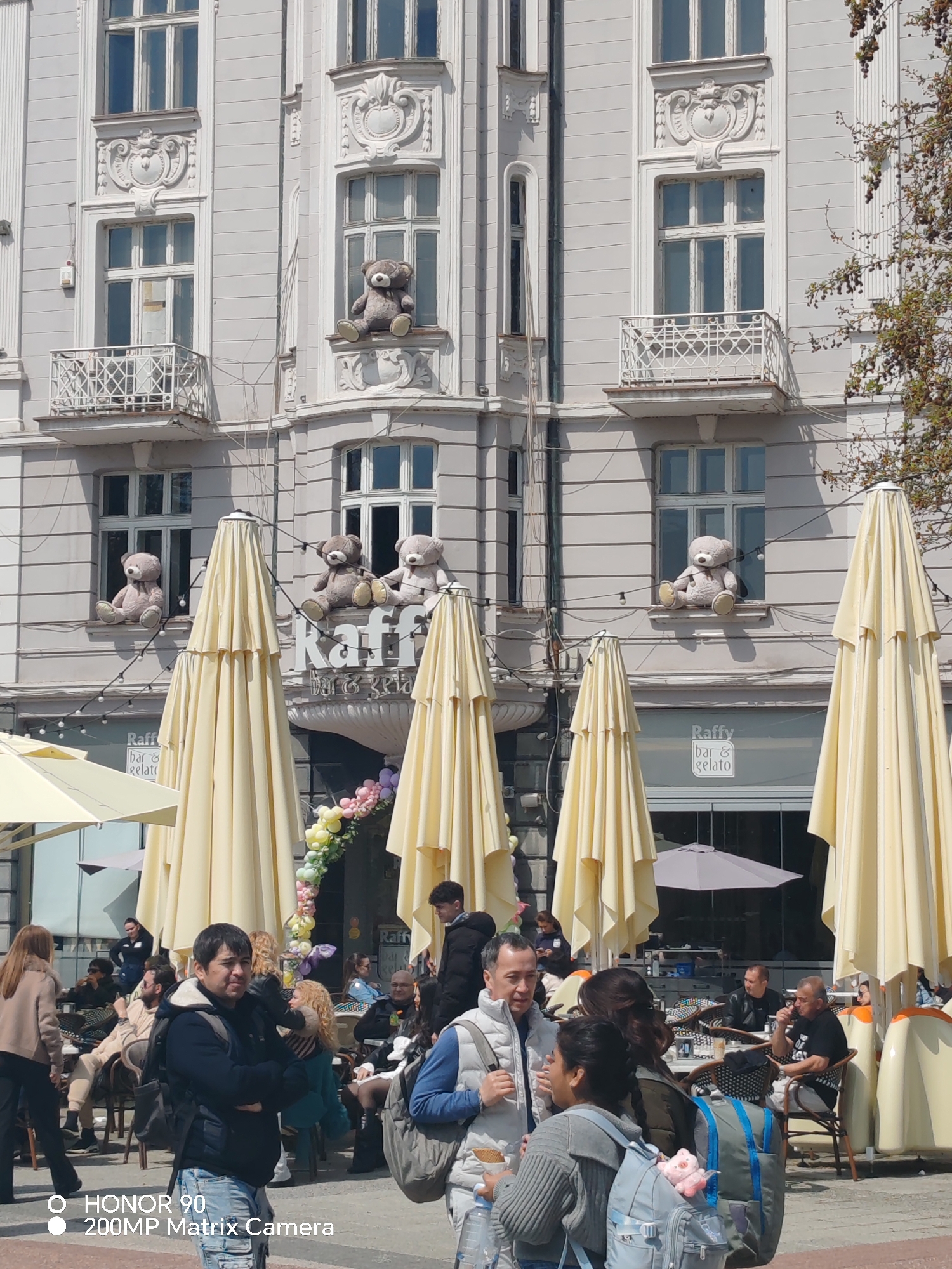 The image depicts a lively street scene in Plovdiv, Bulgaria, in front of a building decorated with large teddy bears on window ledges. The building houses a café or bar called "Raffy bar & gelato." Several large, closed parasols are positioned in front of the building, suggesting an outdoor seating area.
A diverse group of people fills the foreground, walking and interacting with each other. Some are standing and talking, while others are seated at tables under the parasols, seemingly enjoying refreshments. The attire of the people indicates a casual, daytime setting.
The building's facade is grey with white architectural details. The teddy bears, sitting on various windowsills, create a whimsical and playful atmosphere. A floral archway or decoration appears to be located near the entrance of the café.
The overall scene captures a bustling urban environment in Plovdiv on a sunny day.