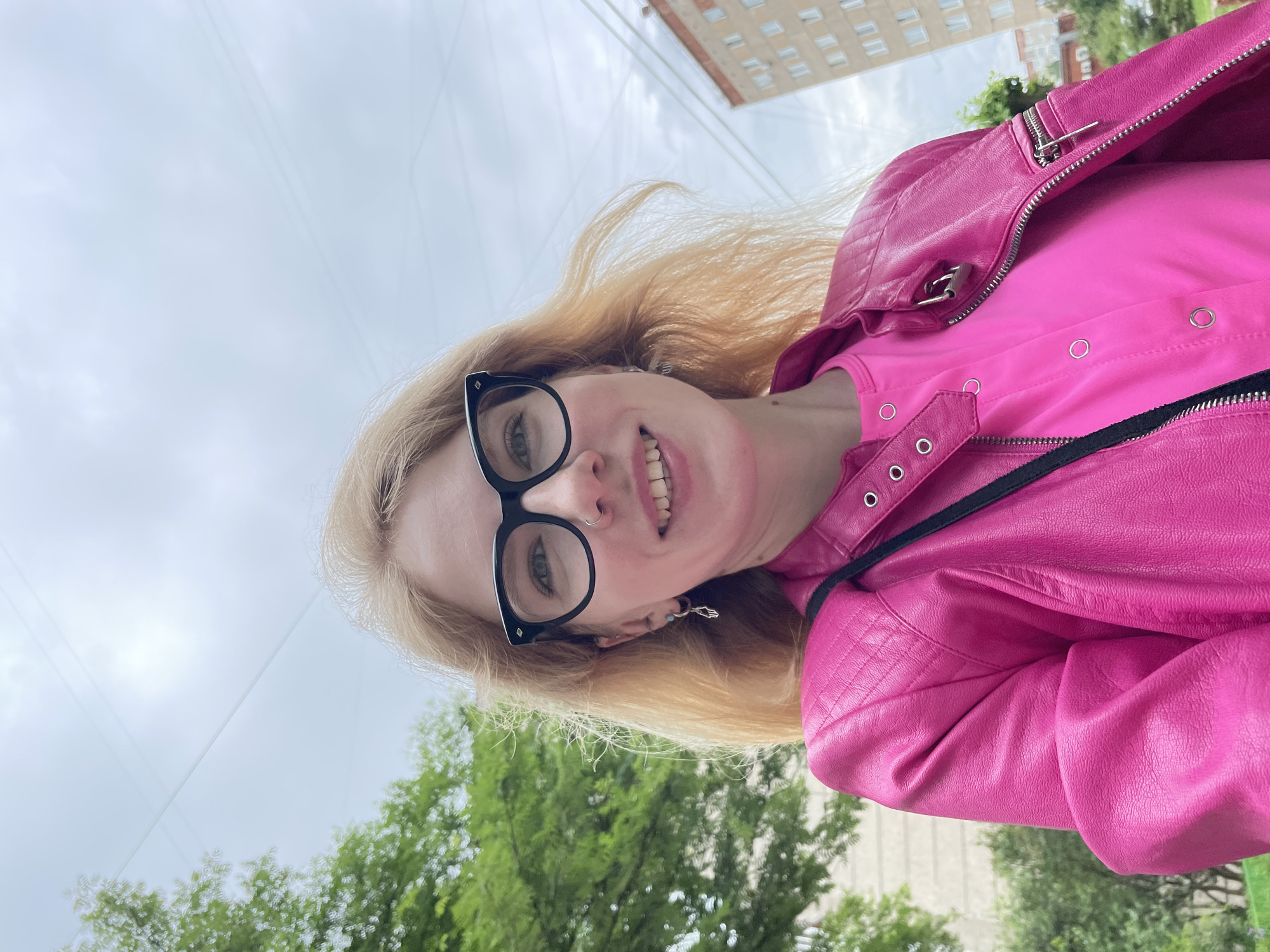 A blonde woman wearing black-rimmed glasses, a nose ring, and earrings poses for a selfie outdoors. She is wearing a bright pink shirt and a pink faux leather jacket with zippers and snaps. Behind her, a green tree is visible on the left, and a building with multiple windows is on the right. The sky is overcast with visible power lines. The context provided states this is a street in Izhevsk, Russia.