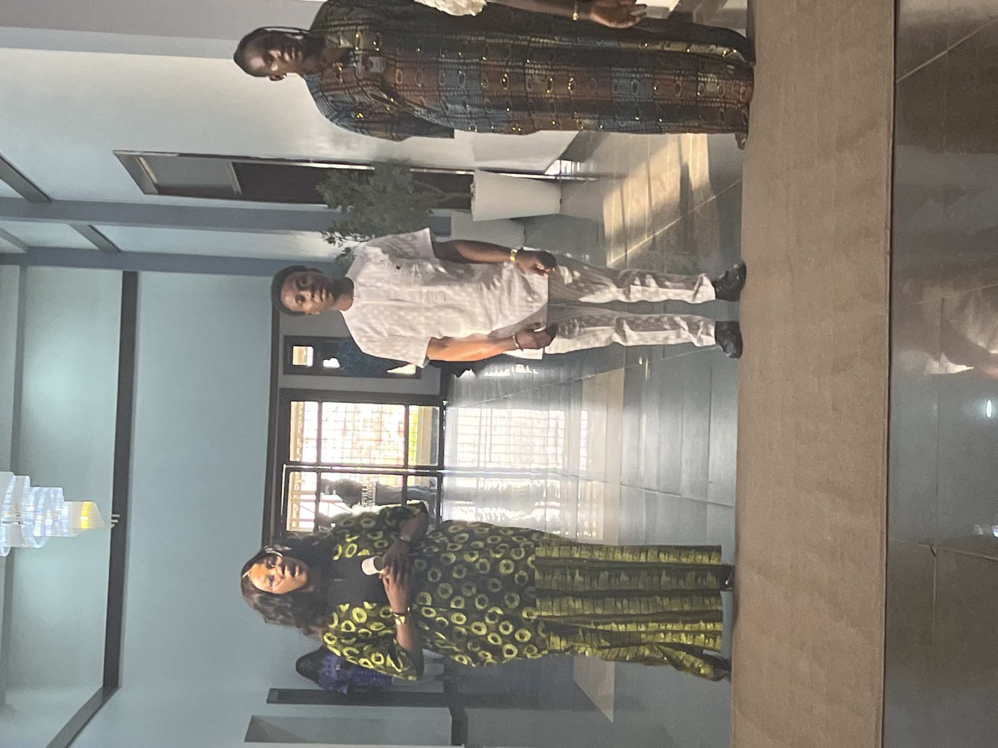 The image shows three people indoors. A woman on the left is wearing a long, patterned green dress. A man in the middle is wearing a white long-sleeved outfit. An older woman on the right is wearing a long, patterned brown dress. They are standing in a building with a view of the outside through a window. The city is Abuja, Nigeria.