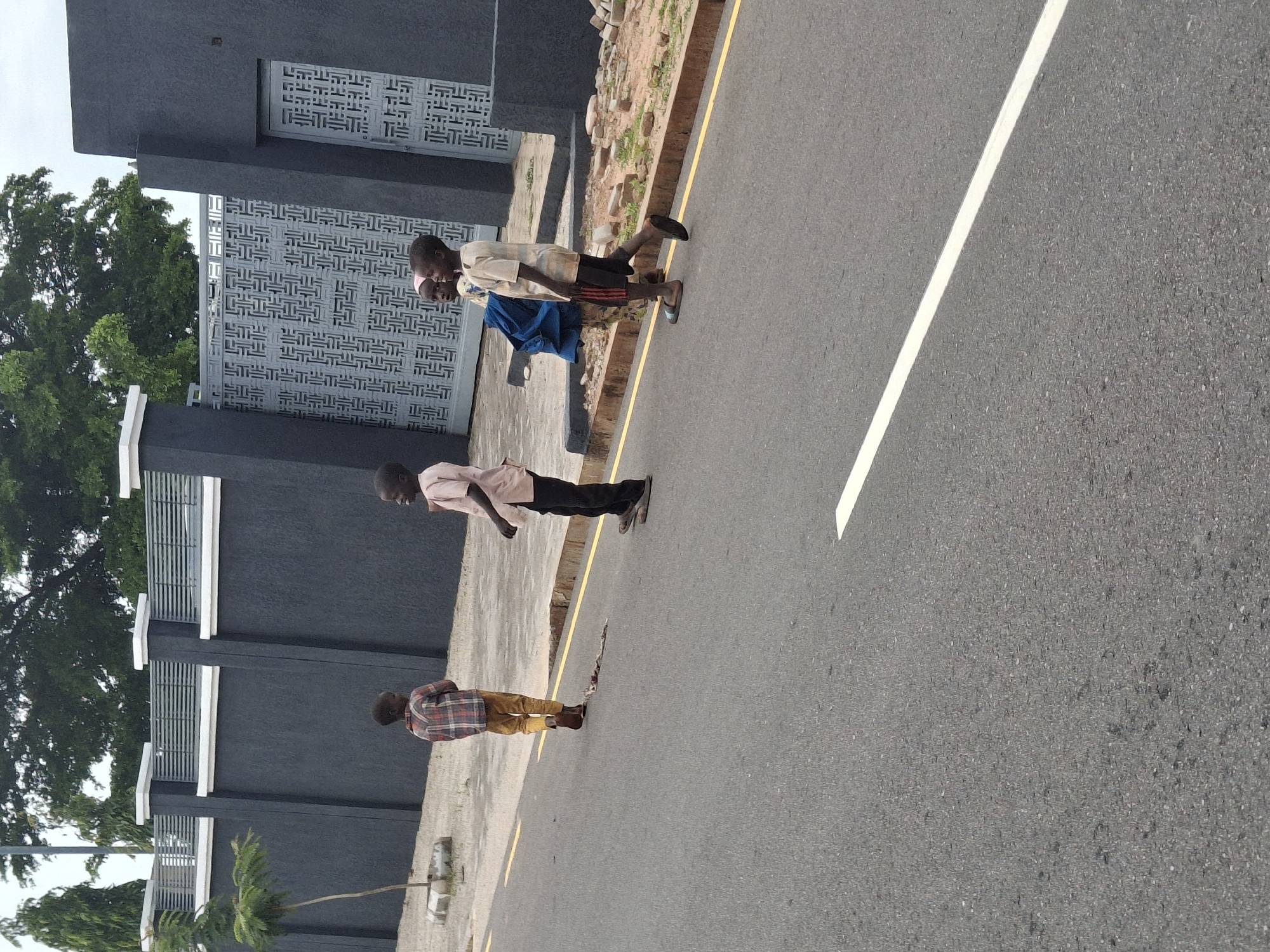 The image depicts a street scene in Gombe, Nigeria, focusing on four children walking on a paved road. The children, appearing to be of school age, are dressed in casual clothing. One child in the foreground on the left is wearing a plaid shirt and yellow trousers. To the right of him, another child in a light pink shirt and black trousers walks with their head turned to the side, seemingly interacting with a third child. This third child, carrying a blue bag, is wearing a light-colored shirt and shorts. A fourth child, walking ahead of them on the right, is wearing a light-colored shirt with red stripes on the sleeves and dark shorts.
The environment suggests an urban setting. A dark gray wall with a decorative metal gate and door forms the backdrop. The wall is topped with a light-colored railing, and a small tree is planted in front of it. The road surface is asphalt with yellow and white lane markings. The overall impression is a mundane street scene with children on their way, potentially to or from school, or simply playing.